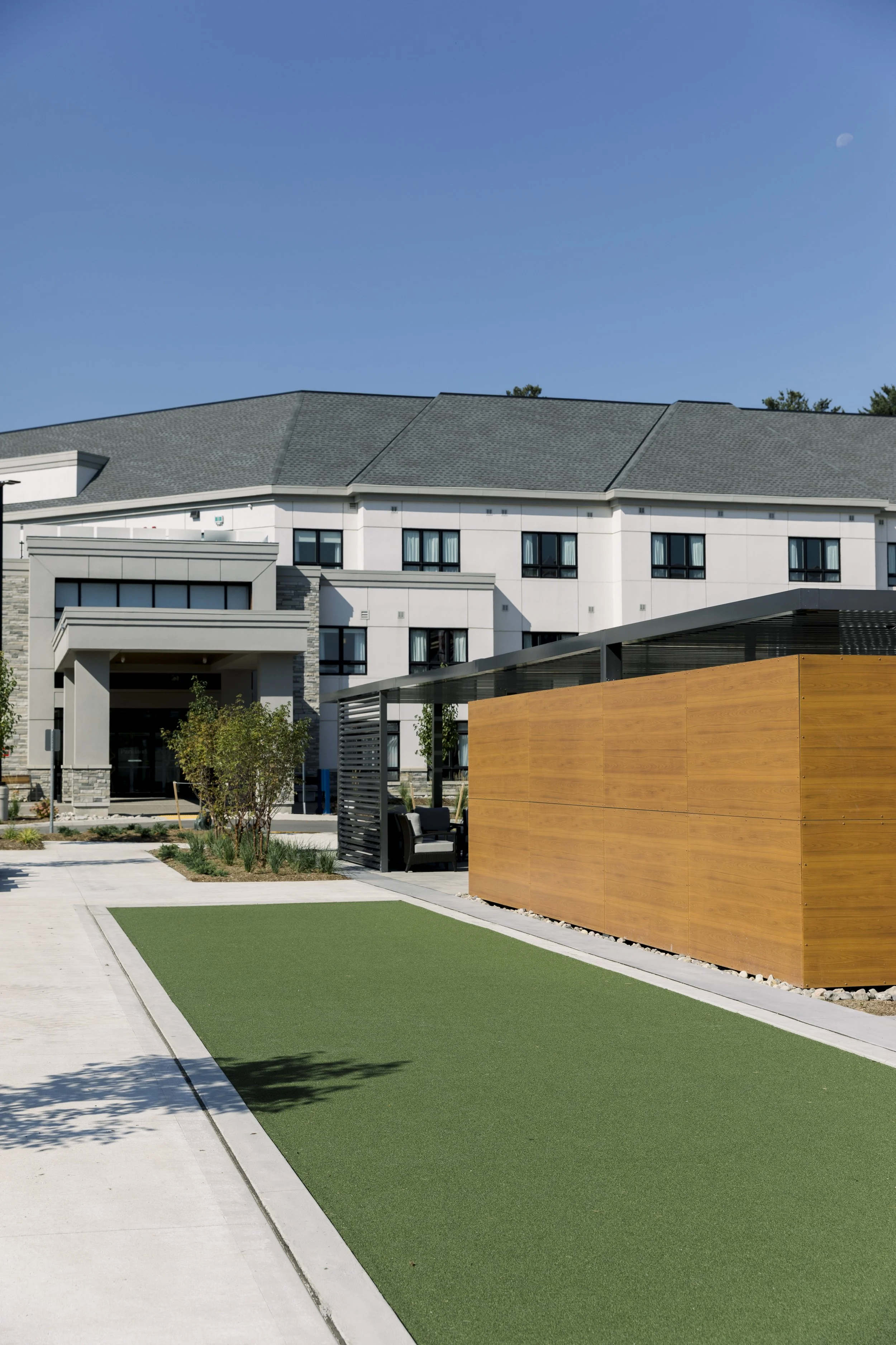 Exterior view of a modern apartment complex with landscaped greenery, a shaded seating area, and a wooden privacy wall under a clear blue sky.