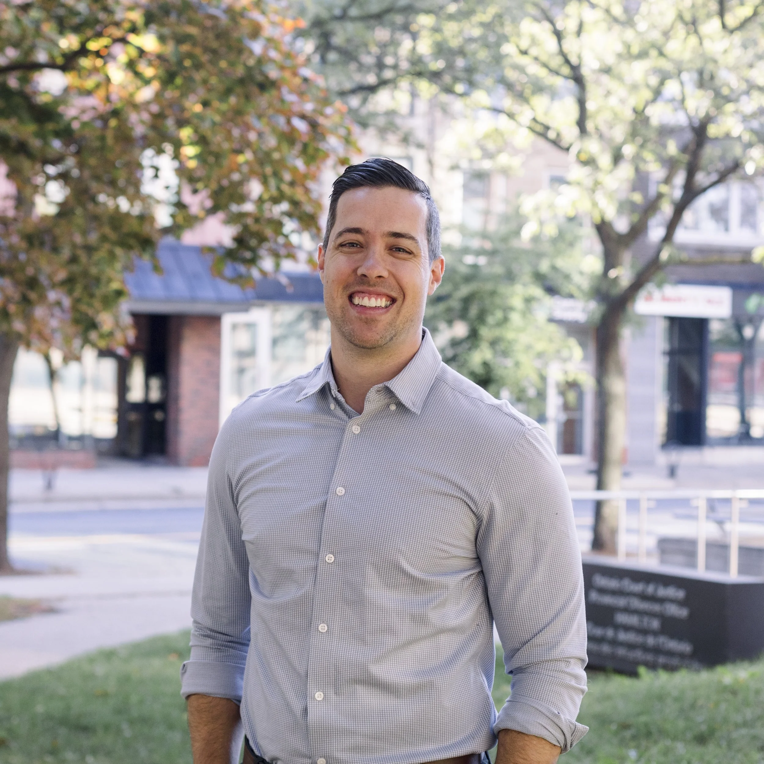 A young man with dark hair, wearing a light gray button-up shirt, smiling outdoors on a sunny day with trees and buildings in the background.