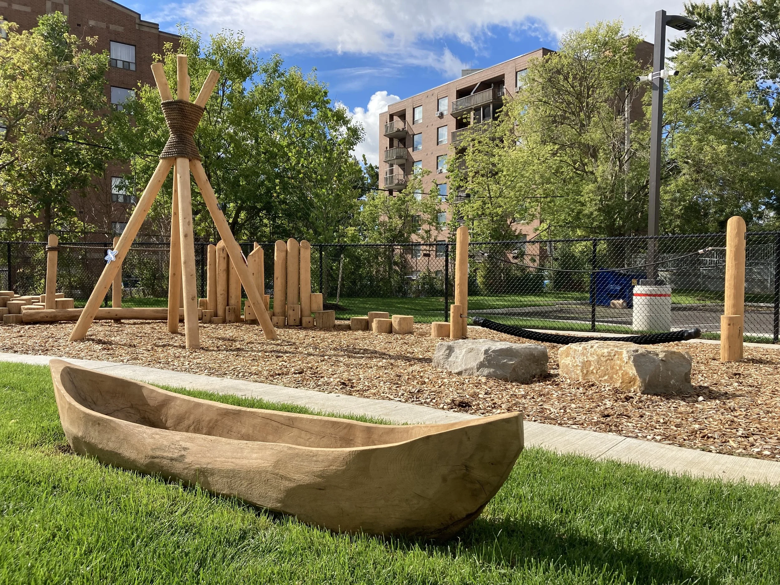 A wooden boat-shaped structure in a green grassy area in a playground, with a wood chip surface and a wooden play set with a rope and logs in the background, surrounded by trees and apartment buildings.