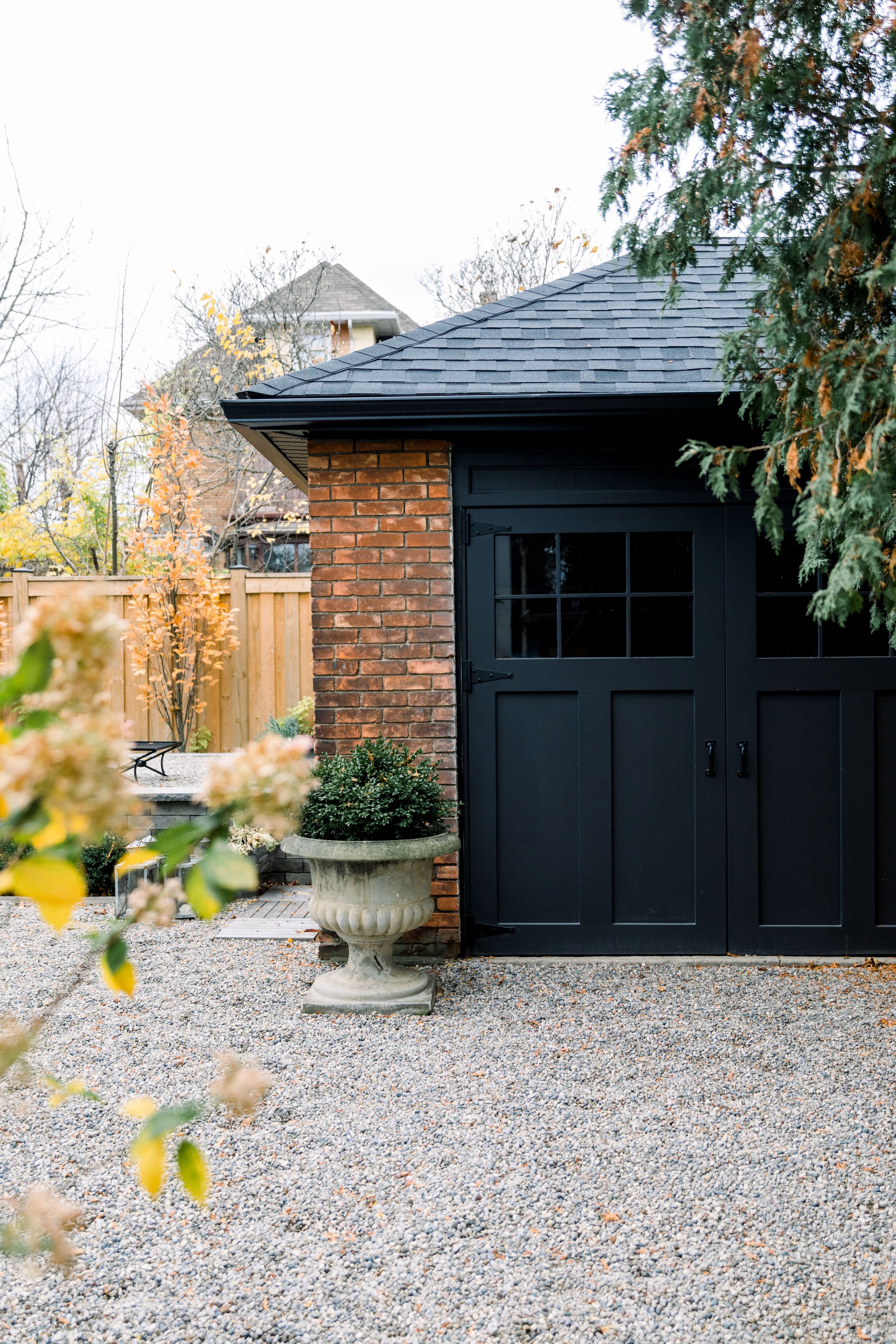 A backyard with a brick wall and a black barn door, gravel ground, and a large decorative planter with greenery, surrounded by trees and a wooden fence.