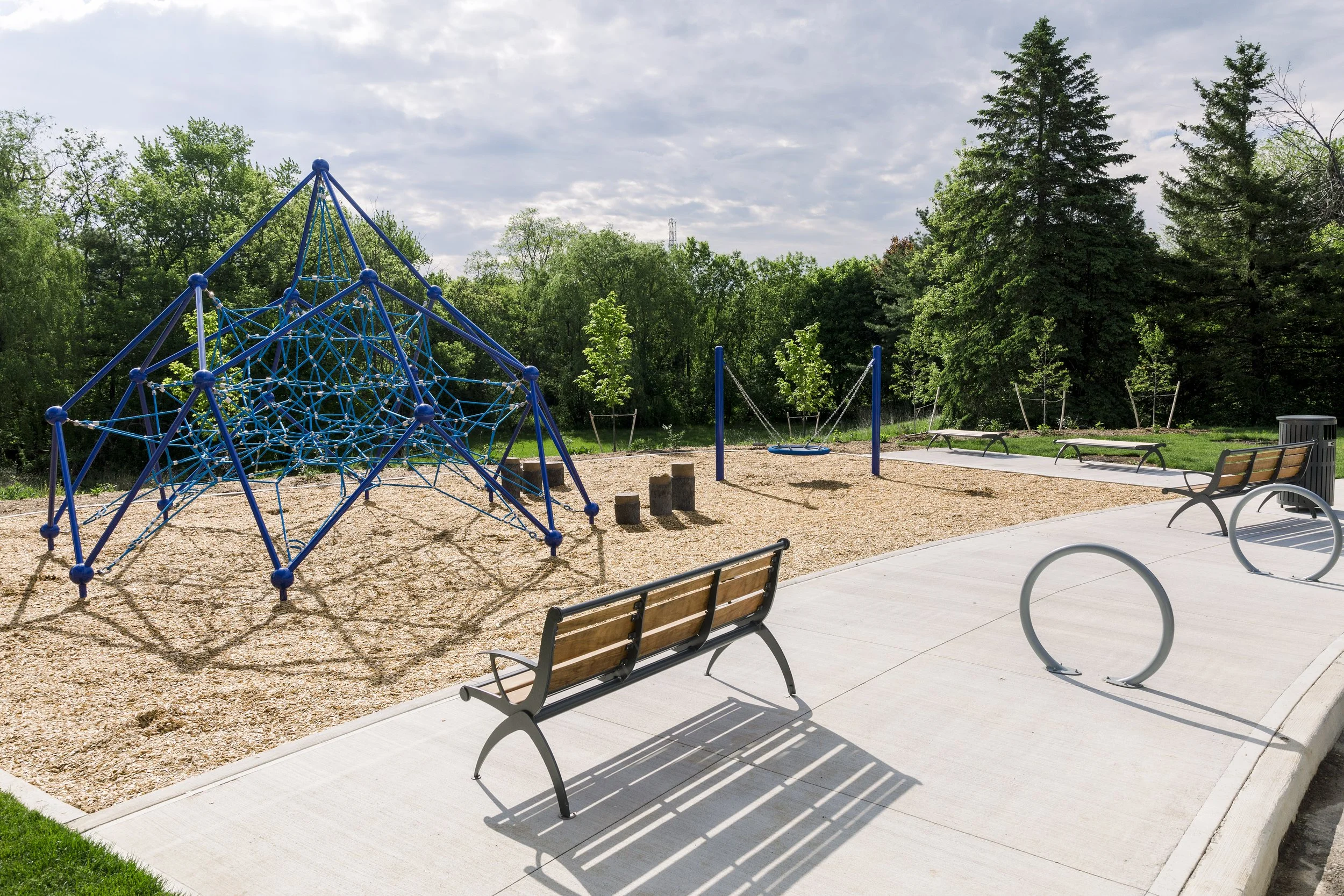 Empty playground with climbing structure, swings, benches, and trees in a park on a sunny day.