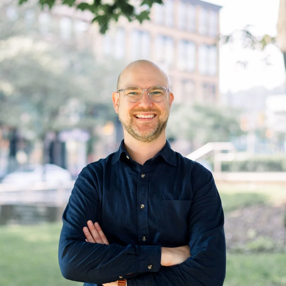 A smiling man with glasses and a beard, wearing a dark blue shirt, standing outdoors with his arms crossed. Blurred trees and buildings are in the background.
