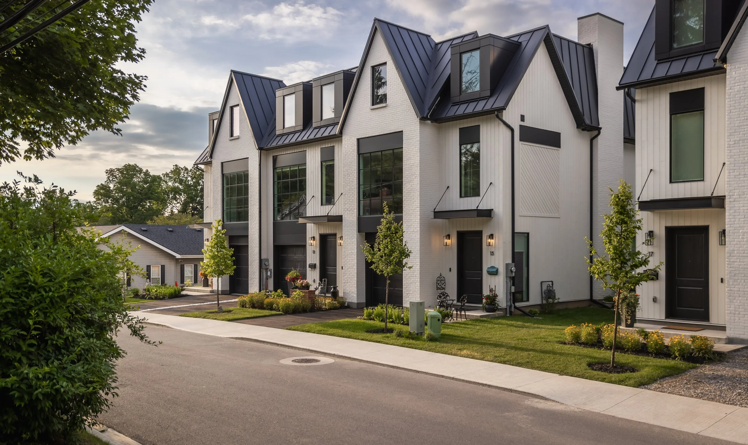 Modern townhouse row with white walls, black accents, and steep metal roofs, featuring front yard with trees and small garden.