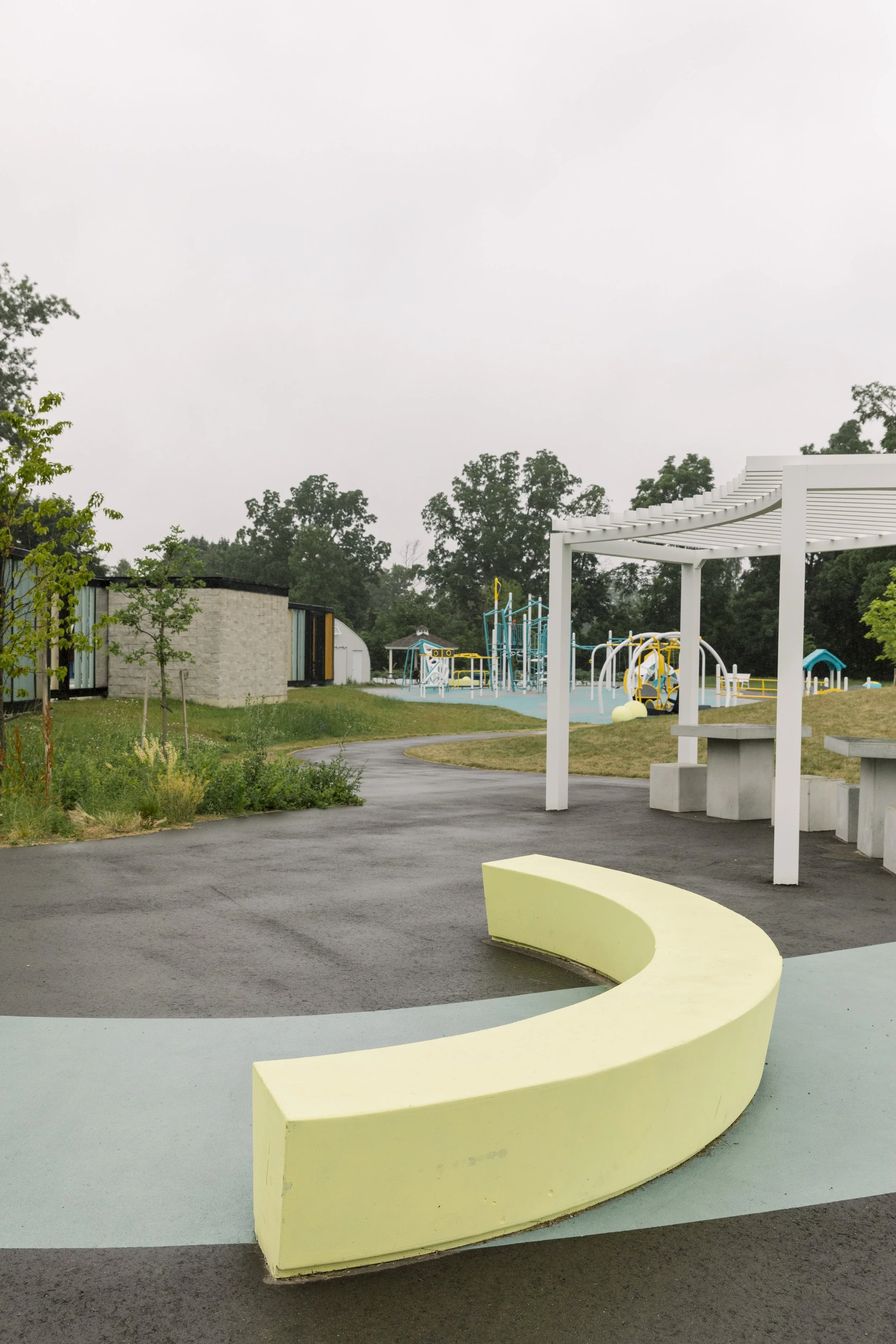 Empty playground area with yellow curved seating, paved paths, grassy areas, a white pergola, and a playground with blue and yellow equipment in the background under cloudy sky.