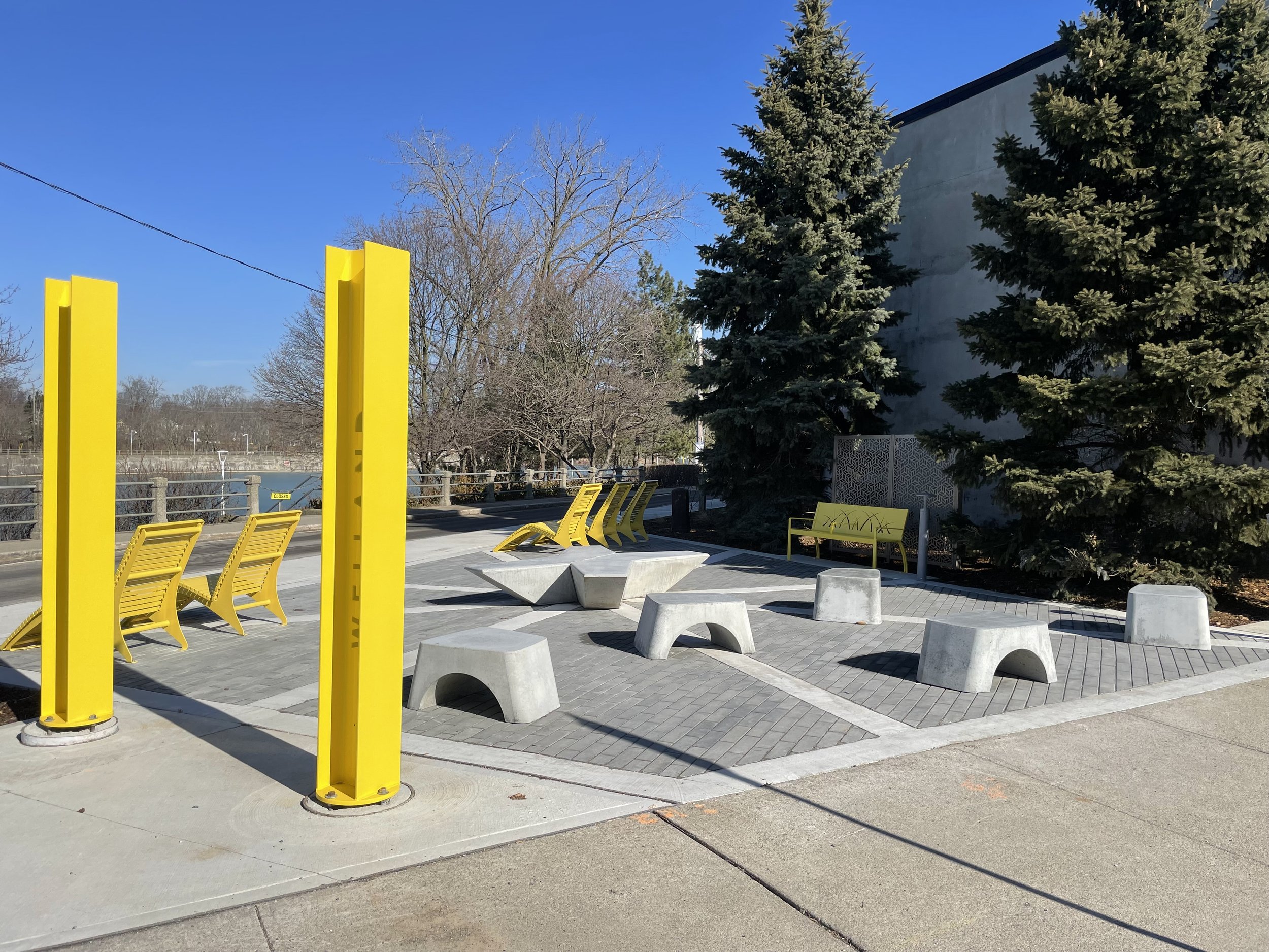 Outdoor seating area with yellow benches, modern concrete stools and tables, and yellow vertical structures in a park-like setting with trees and a creek in the background.