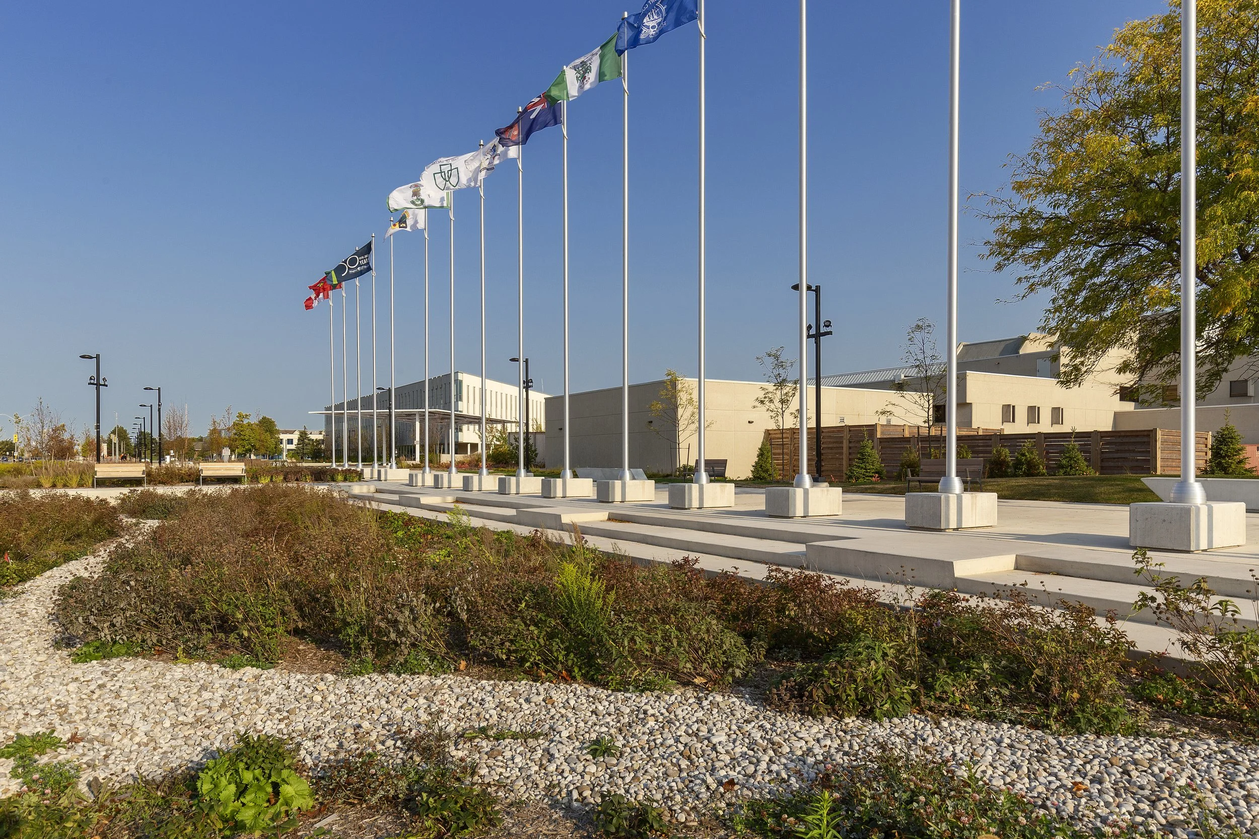 A row of flags on tall flagpoles outside a modern building, with a bright blue sky and landscaped garden in the foreground.