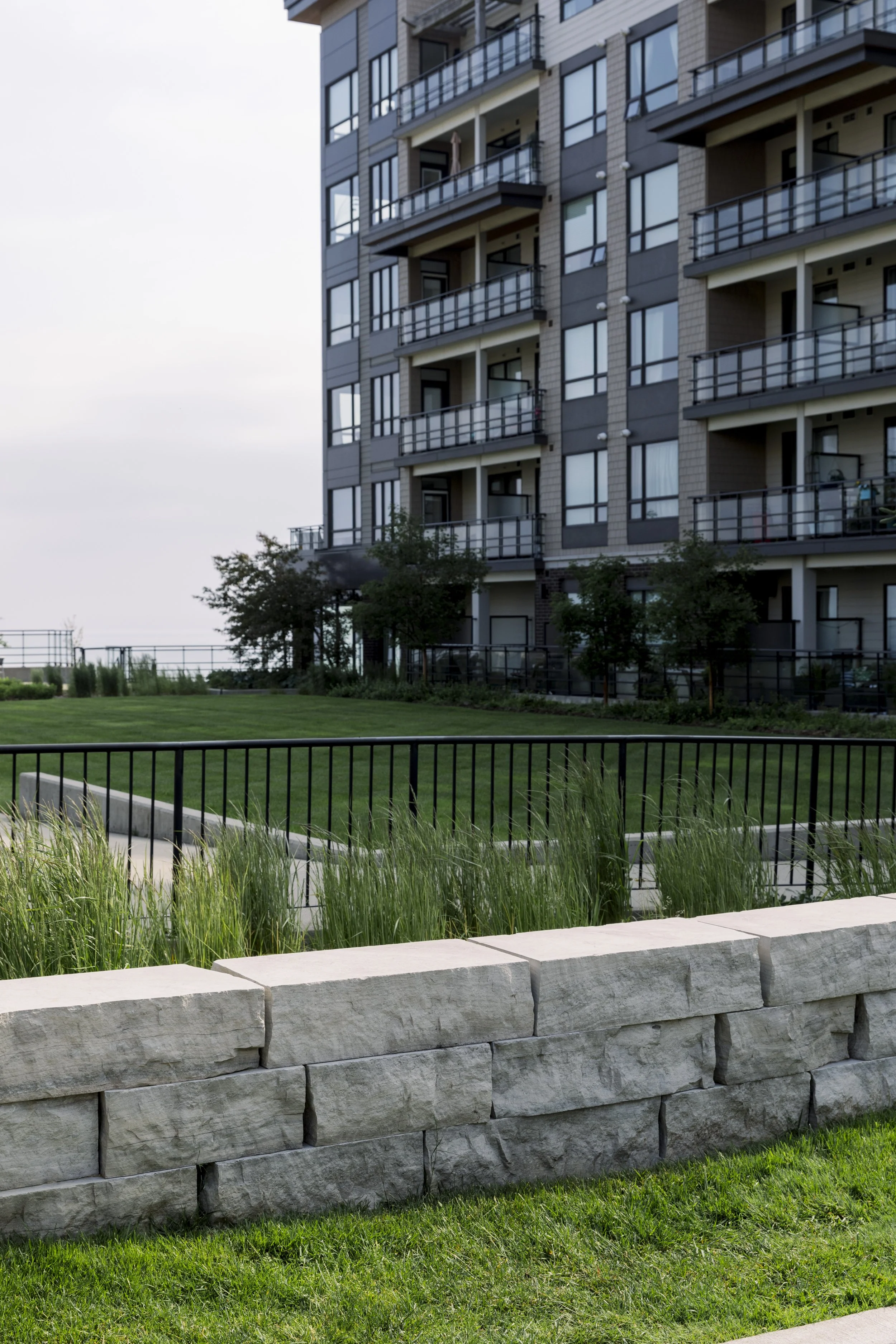 View of a modern apartment building with balconies, a well-maintained lawn, and a stone-edged garden bed with green grass and plants.