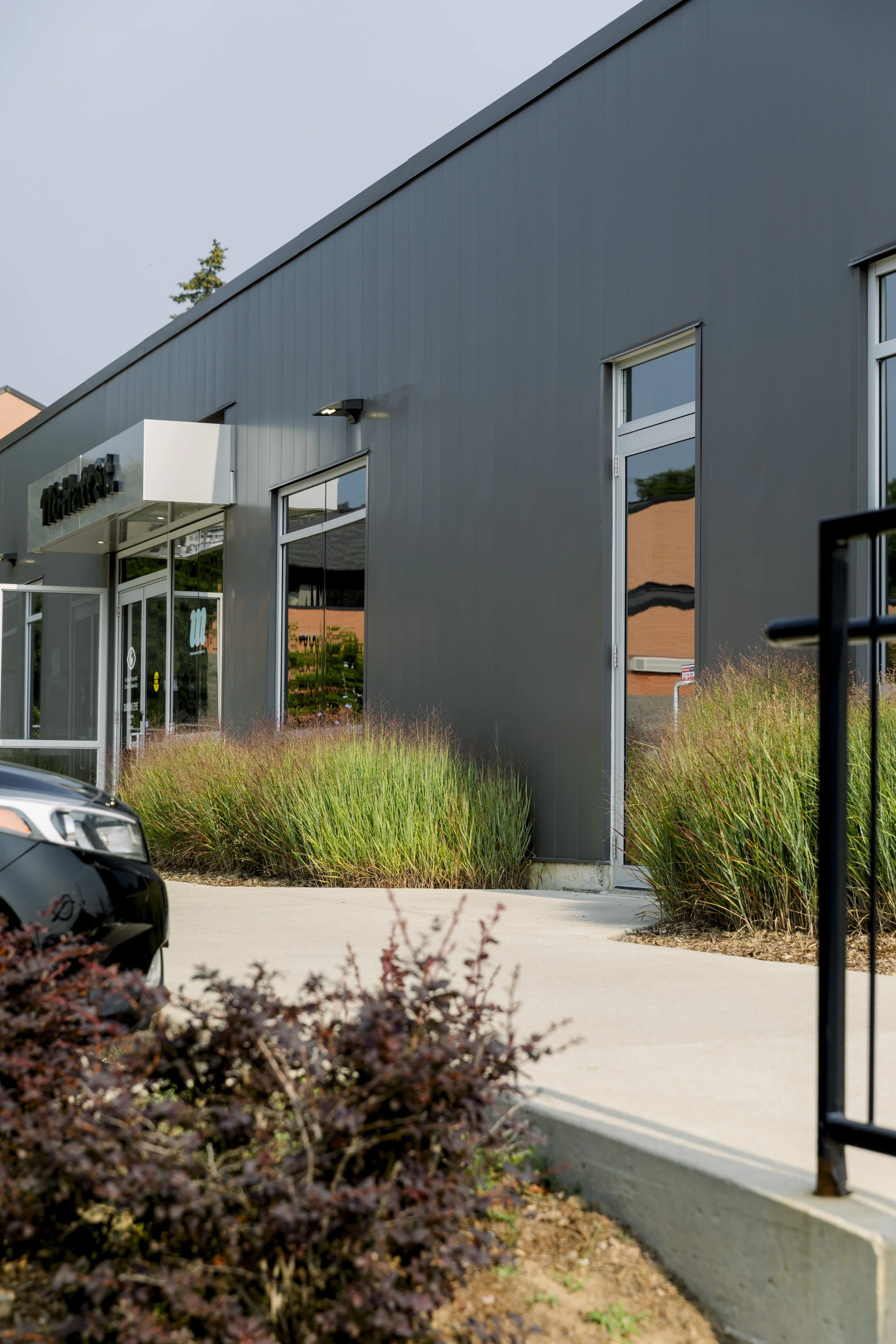 Modern commercial building with dark gray metal siding, glass entrance doors, large windows, surrounded by landscaped bushes and plants, with a concrete sidewalk and a nearby parked car.