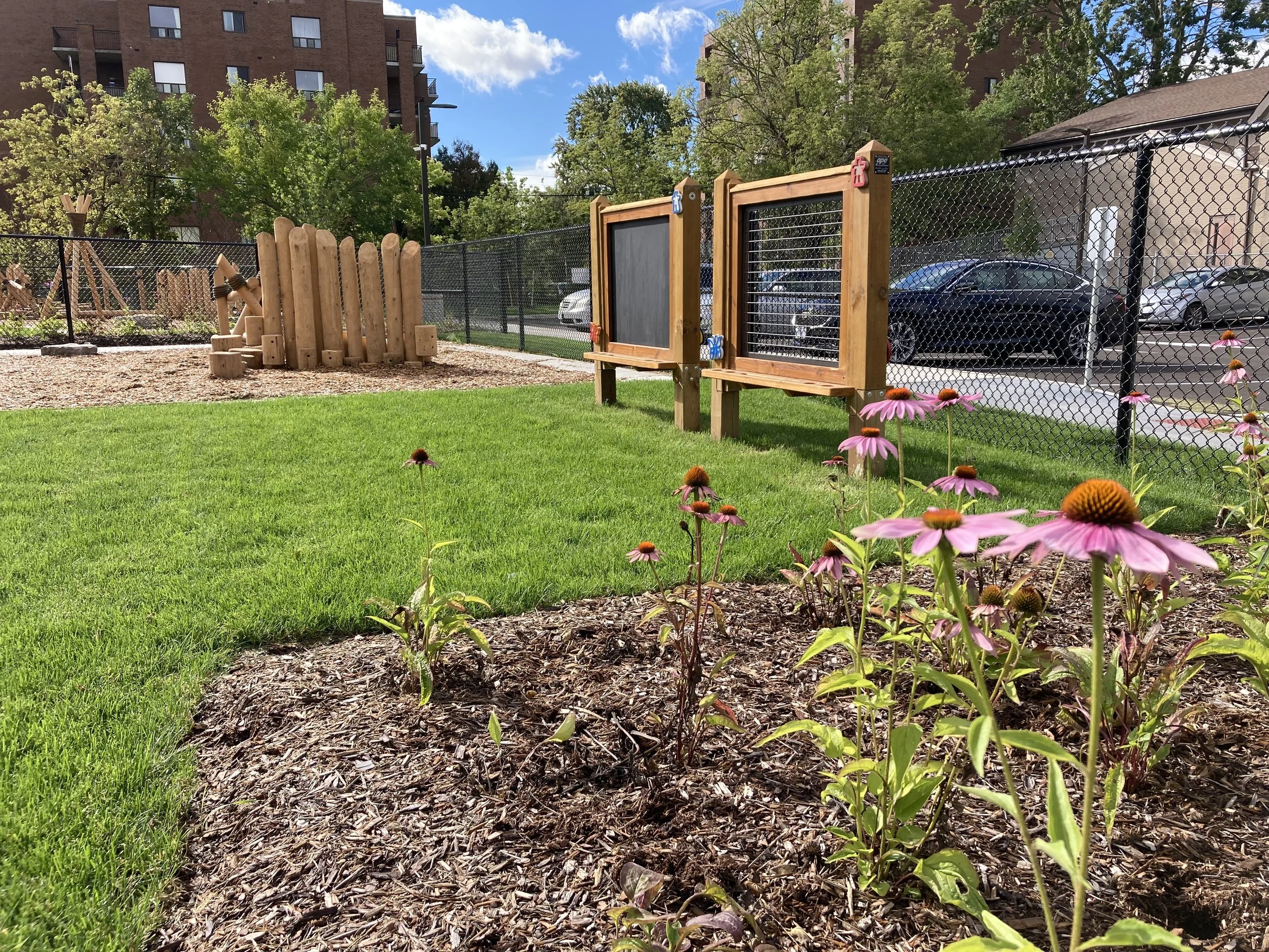A small fenced playground with a grassy area, pink flowers in the foreground, wooden structures including two blackboards or chalkboards, and a wooden climbing frame with a slide in the background. There are trees, a parking lot with cars, and an apa