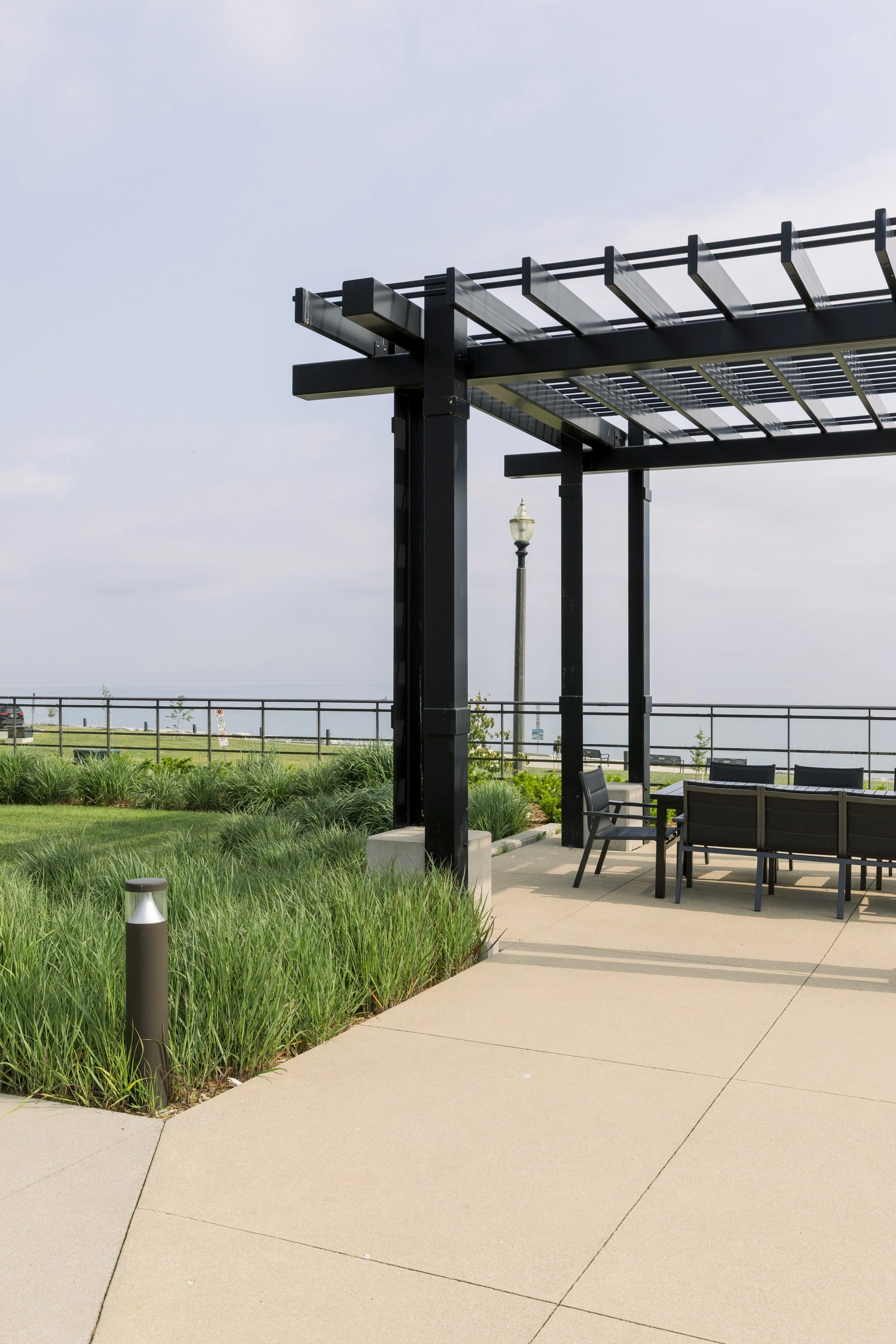 An outdoor park area with a black pergola, green bushes, benches, a lamp post, and a paved walkway.