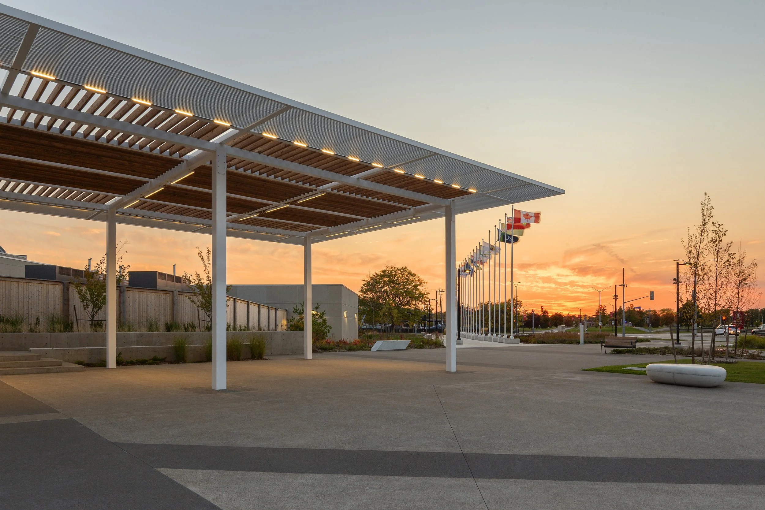 Empty outdoor public space at sunset with modern pavilion structure, flagpoles with flags, benches, trees, and streetlights.