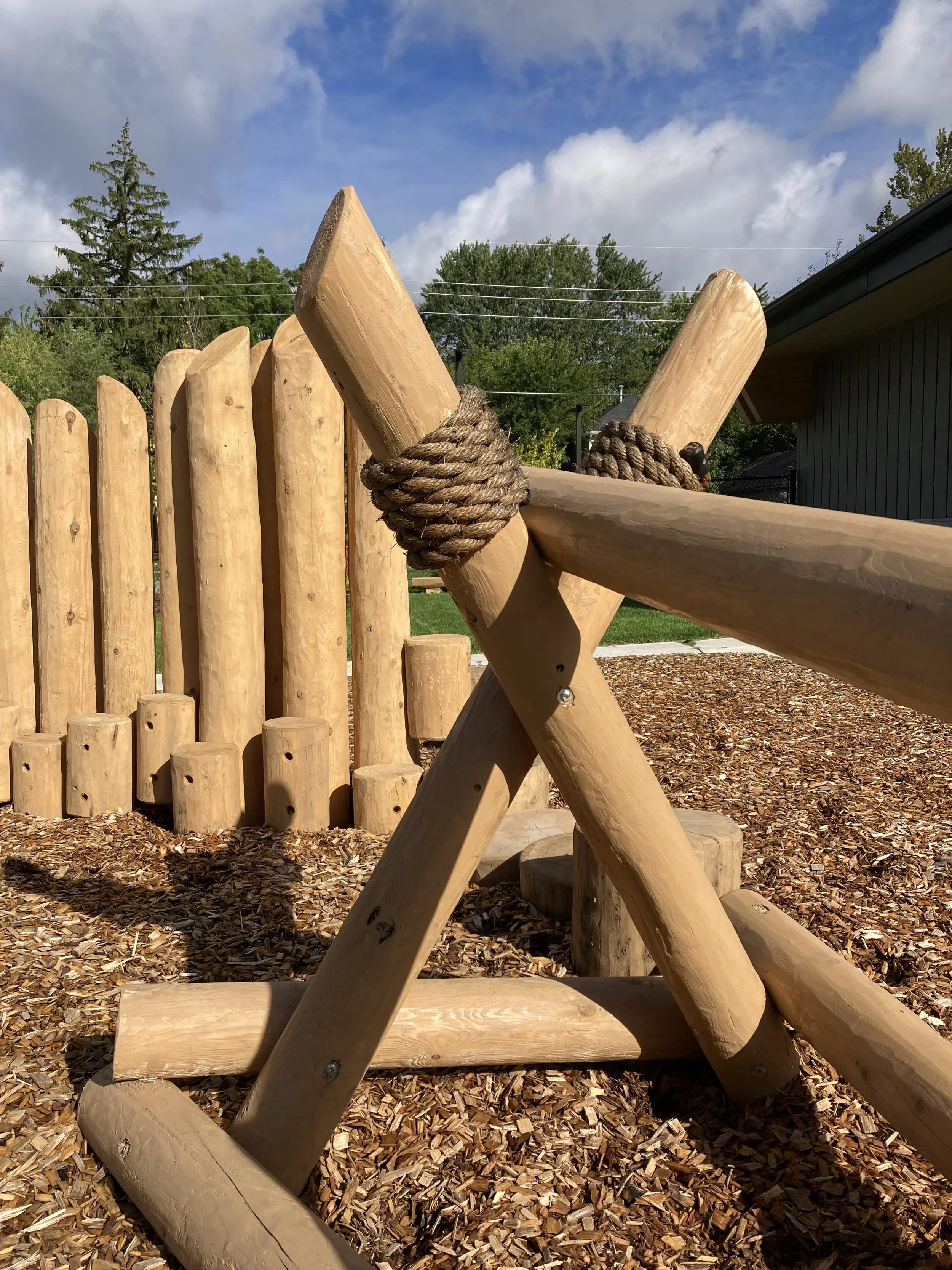 Close-up of wooden logs and ropes in a playground structure with a wood chip ground cover, fence, trees, and a building in the background.