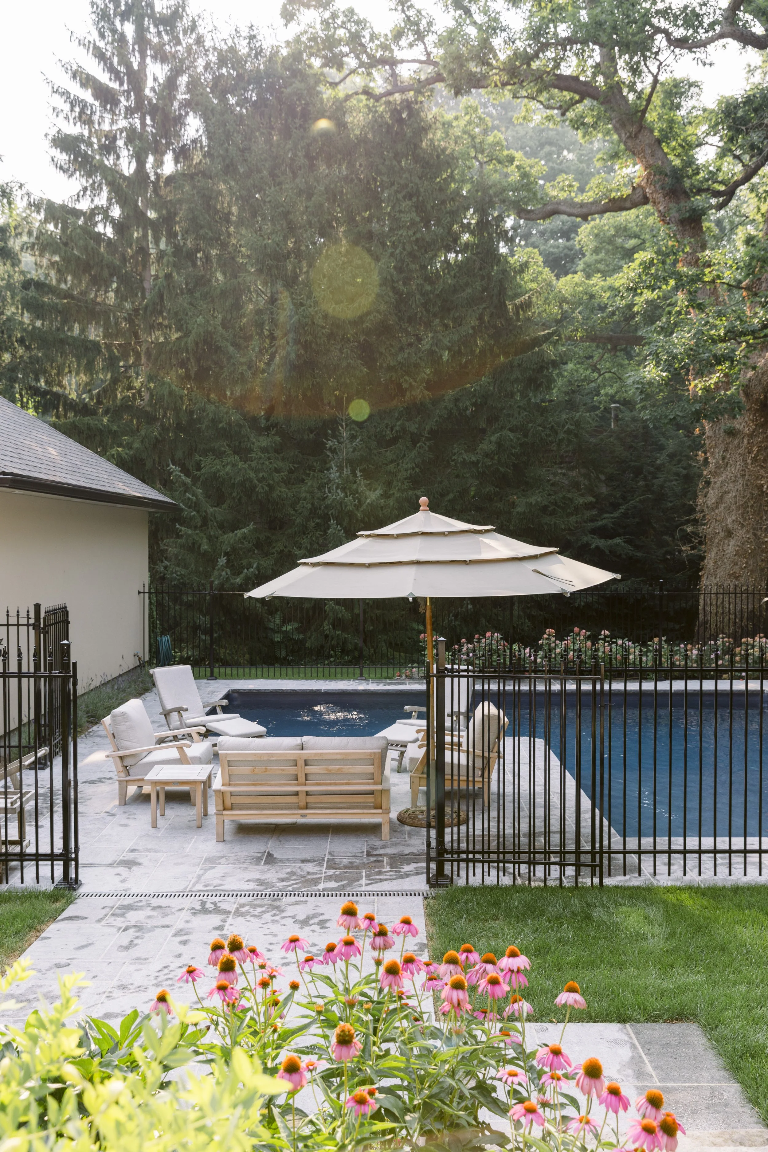 Residential backyard with swimming pool surrounded by a black metal fence, patio furniture, and a large umbrella. There are pink flowers in the foreground and trees in the background.