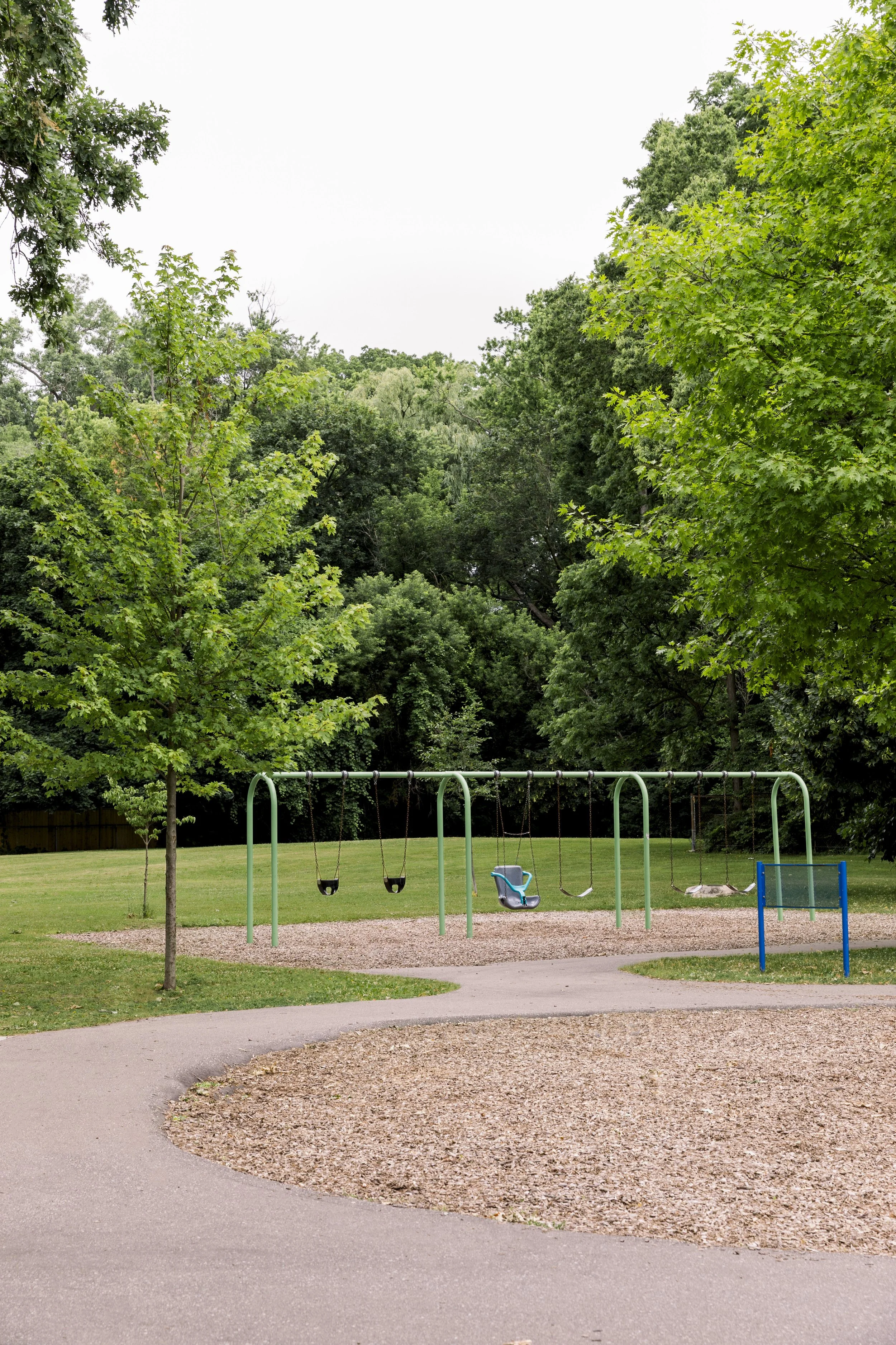 Empty swings and slide in a park surrounded by green trees.