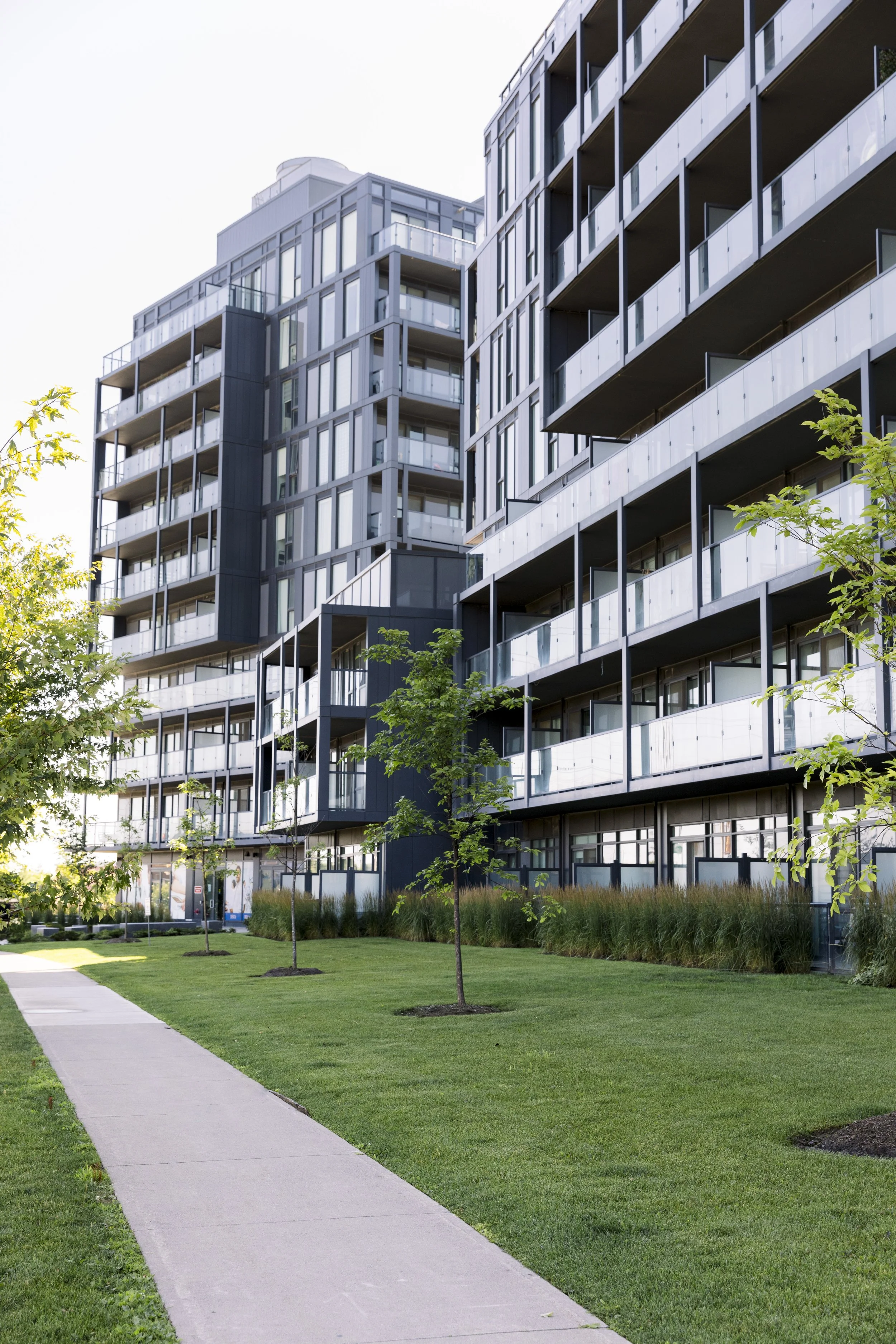 Modern apartment building with glass balconies, surrounded by a well-maintained grassy area and young trees, under a clear sky.