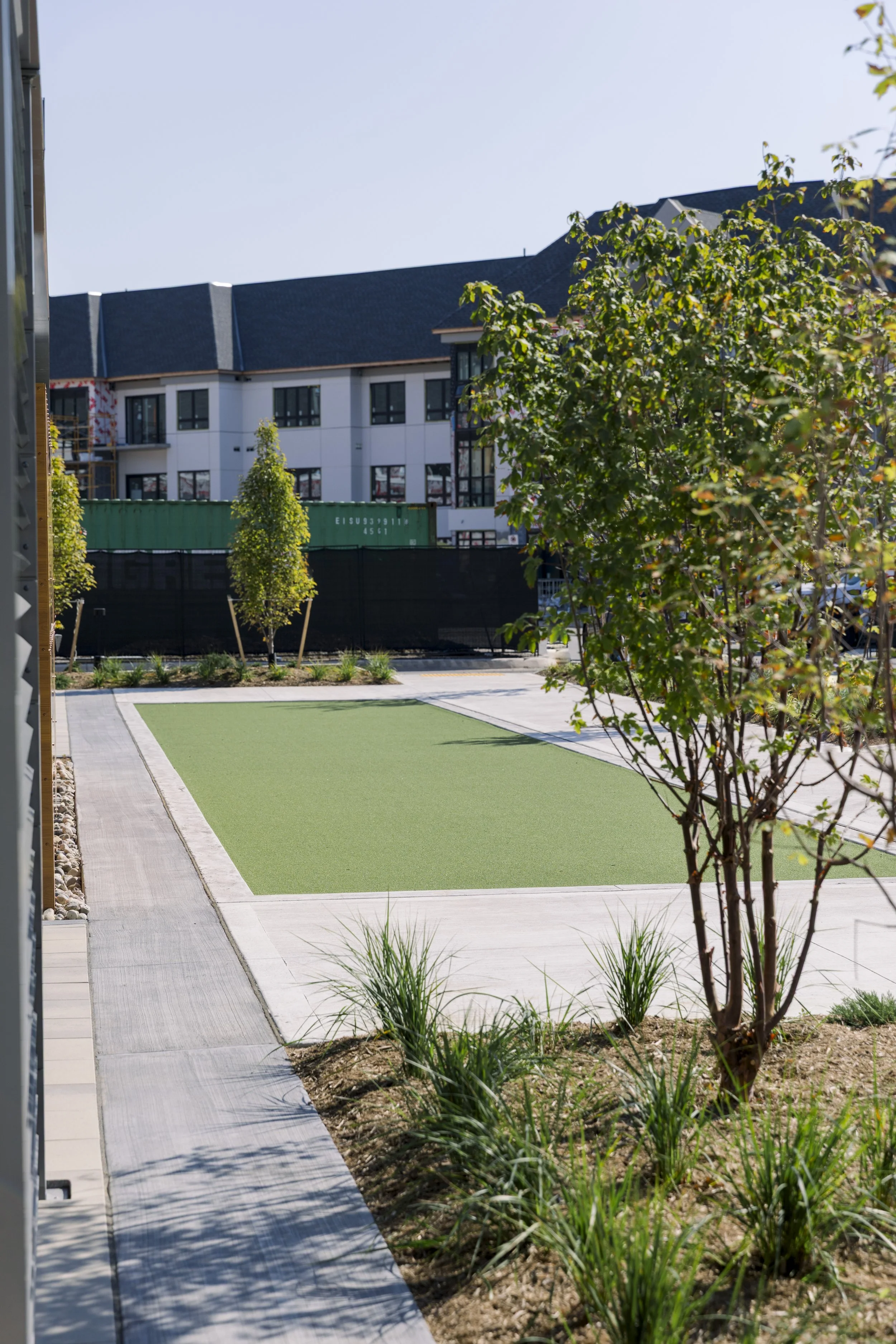 Modern urban courtyard with small trees, shrubs, and a patch of artificial grass, surrounded by paved walkways and a multi-story building in the background.