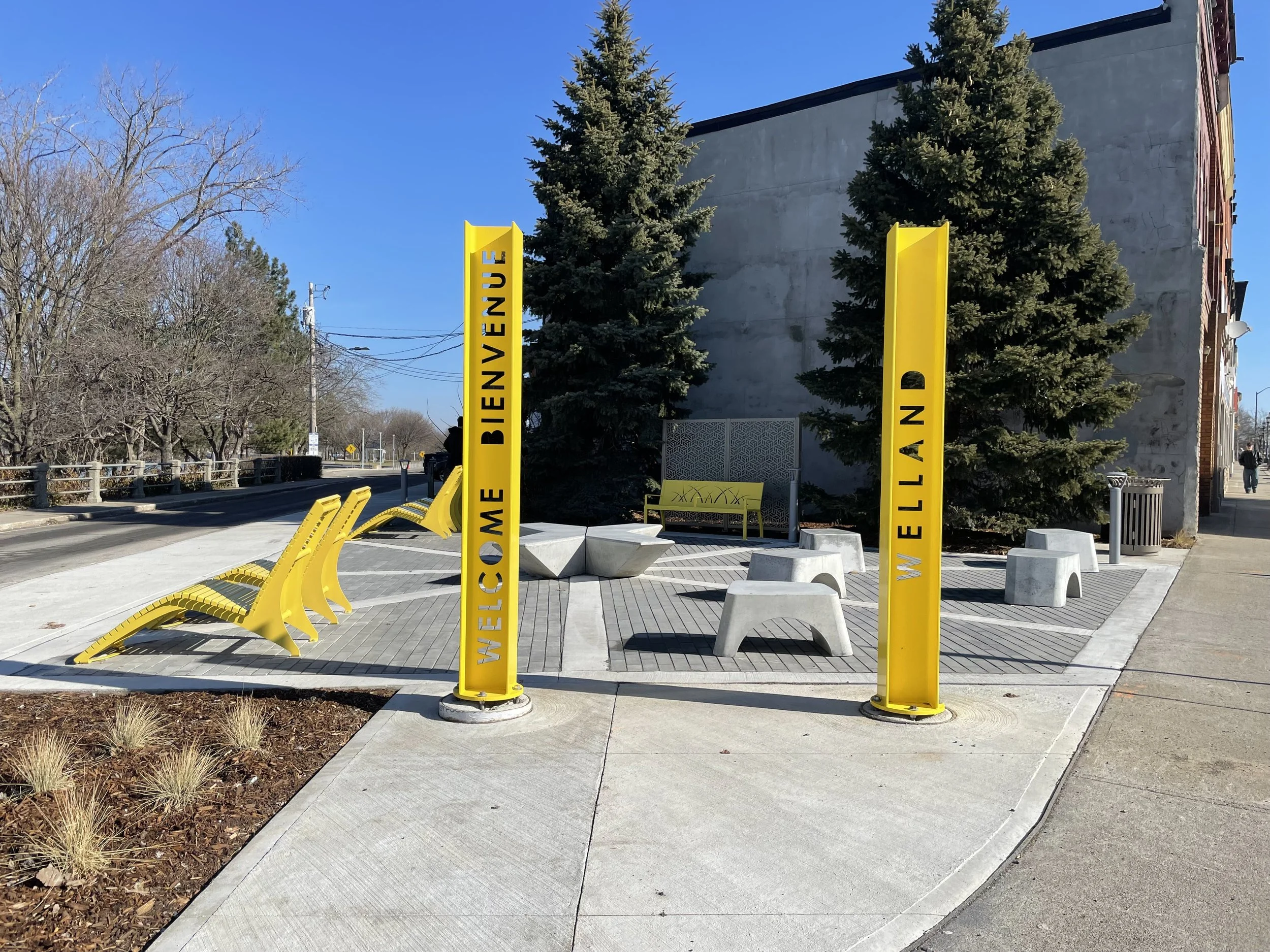 Sidewalk with bright yellow benches and two yellow vertical signs reading "Welcome Bienvenue" and "Welcome Elland" in front of trees and a building with a gray wall, along a street under a blue sky.