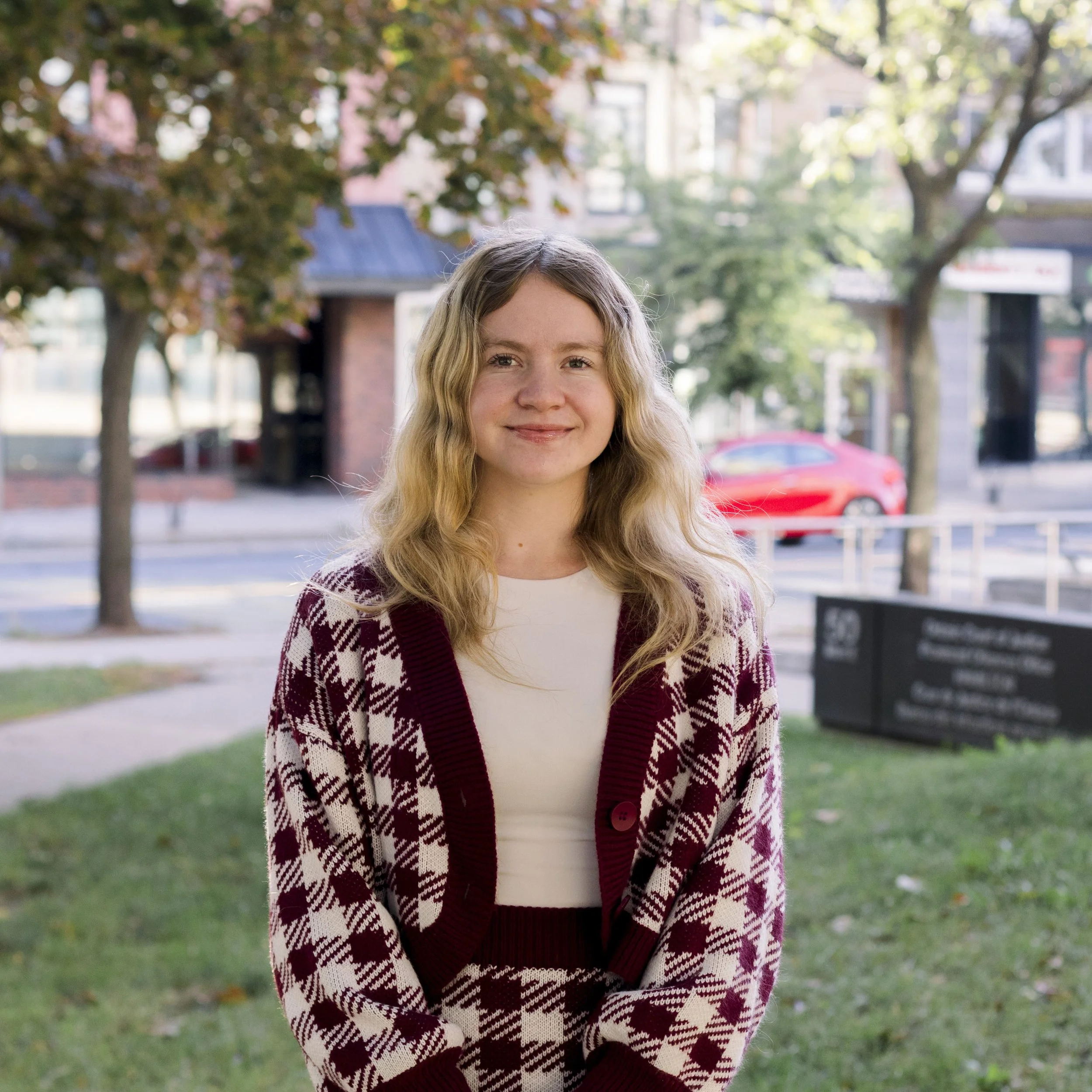 A young woman with wavy blonde hair, wearing a beige top and a red and white plaid cardigan, standing outdoors on a grassy area with trees and buildings in the background.