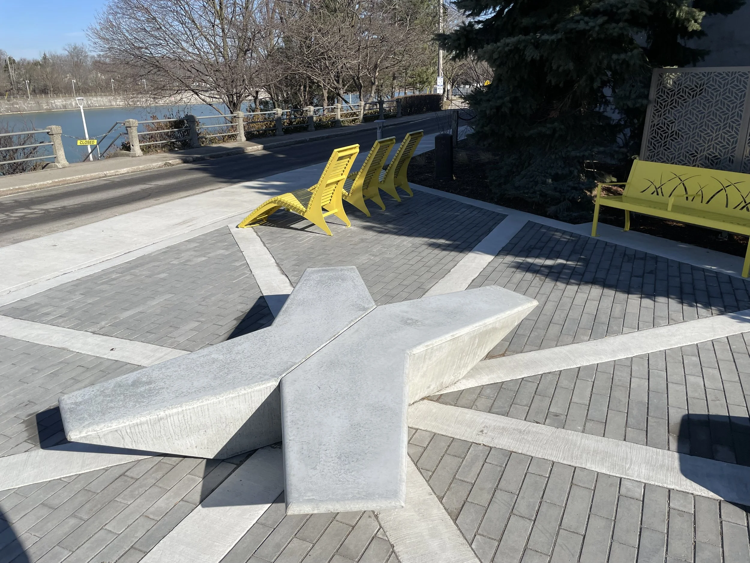 Urban outdoor space with modern concrete benches, yellow chairs, and a yellow bench. The area has patterned pavement and is next to a body of water with a railing and a sign indicating it is closed.