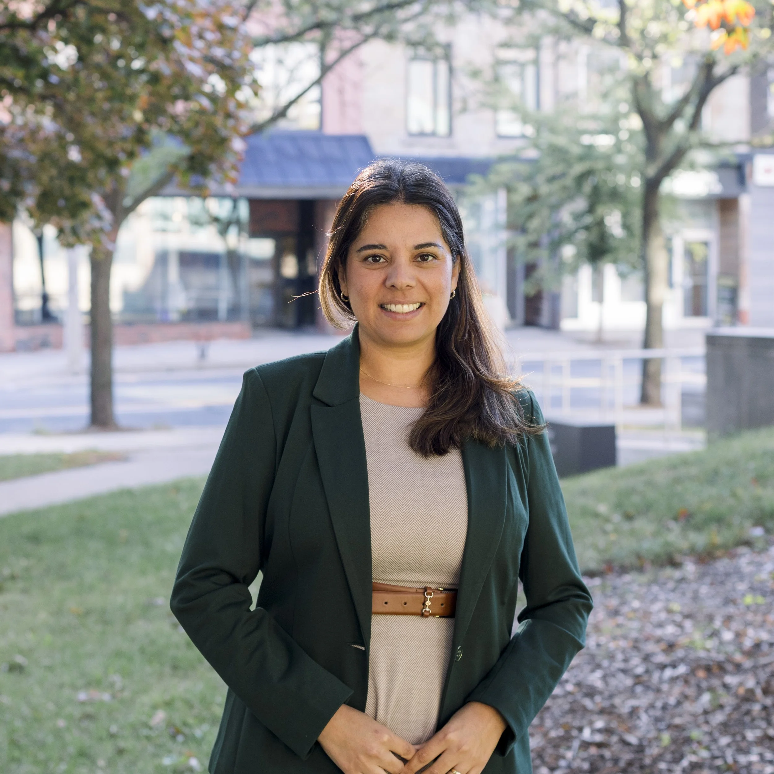 A woman standing outdoors on a sidewalk in a city park with trees and buildings in the background, smiling and looking at the camera.