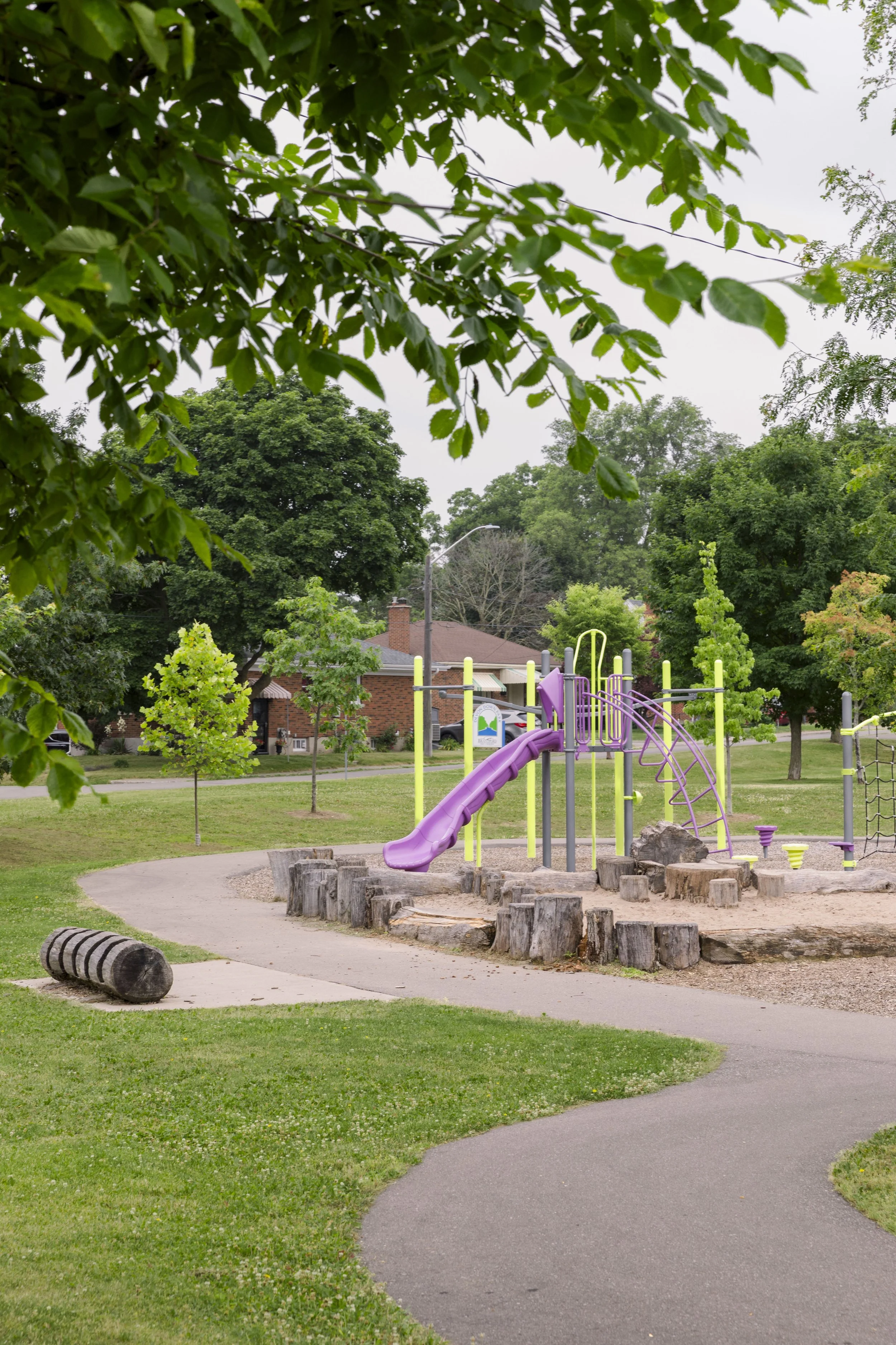 A park playground with a purple slide, climbing structure, and wooden stumps, surrounded by green grass and trees on an overcast day.