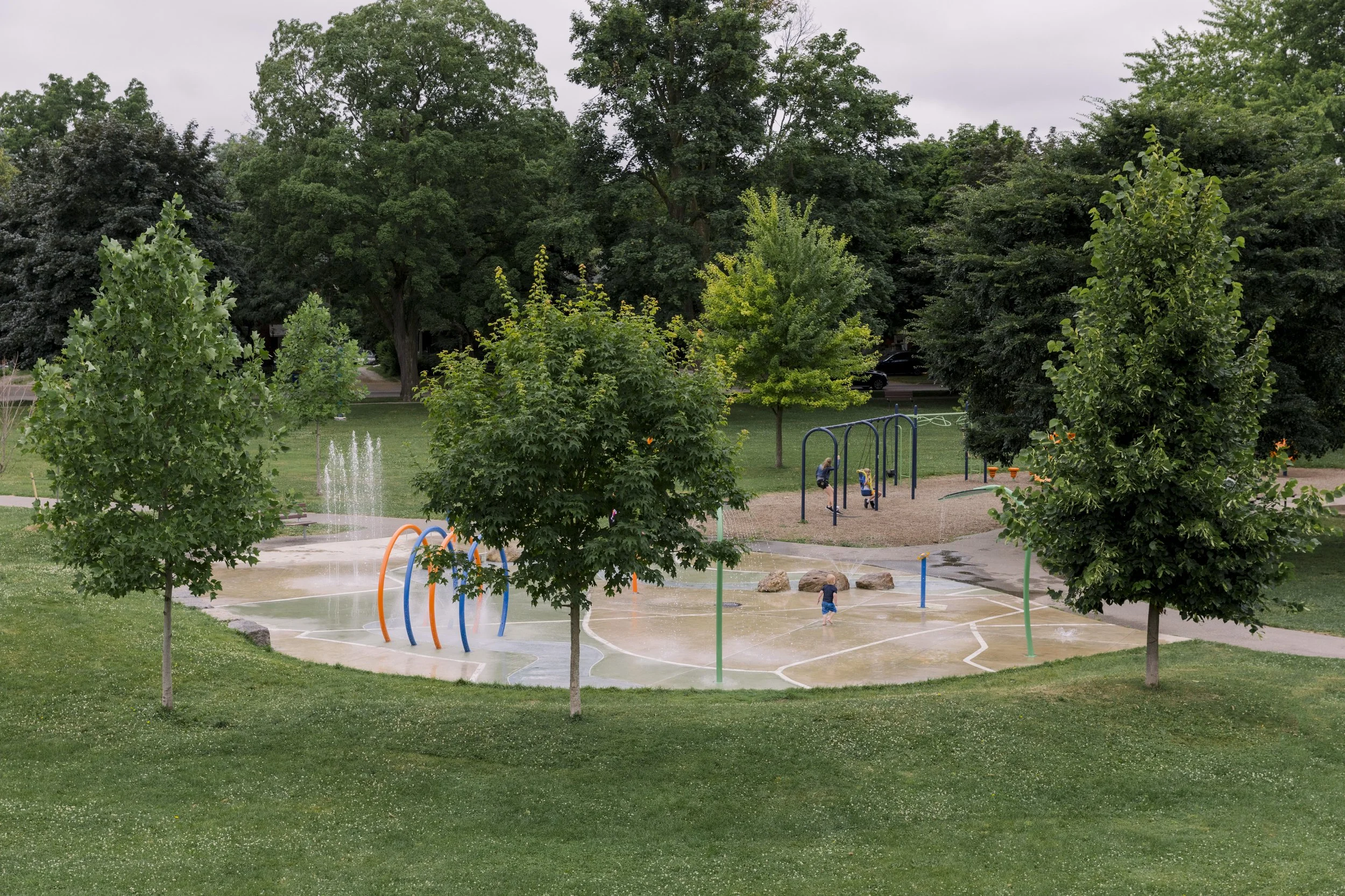 A park with a splash pad and children playing in the water, surrounded by green grass, trees, and playground equipment.
