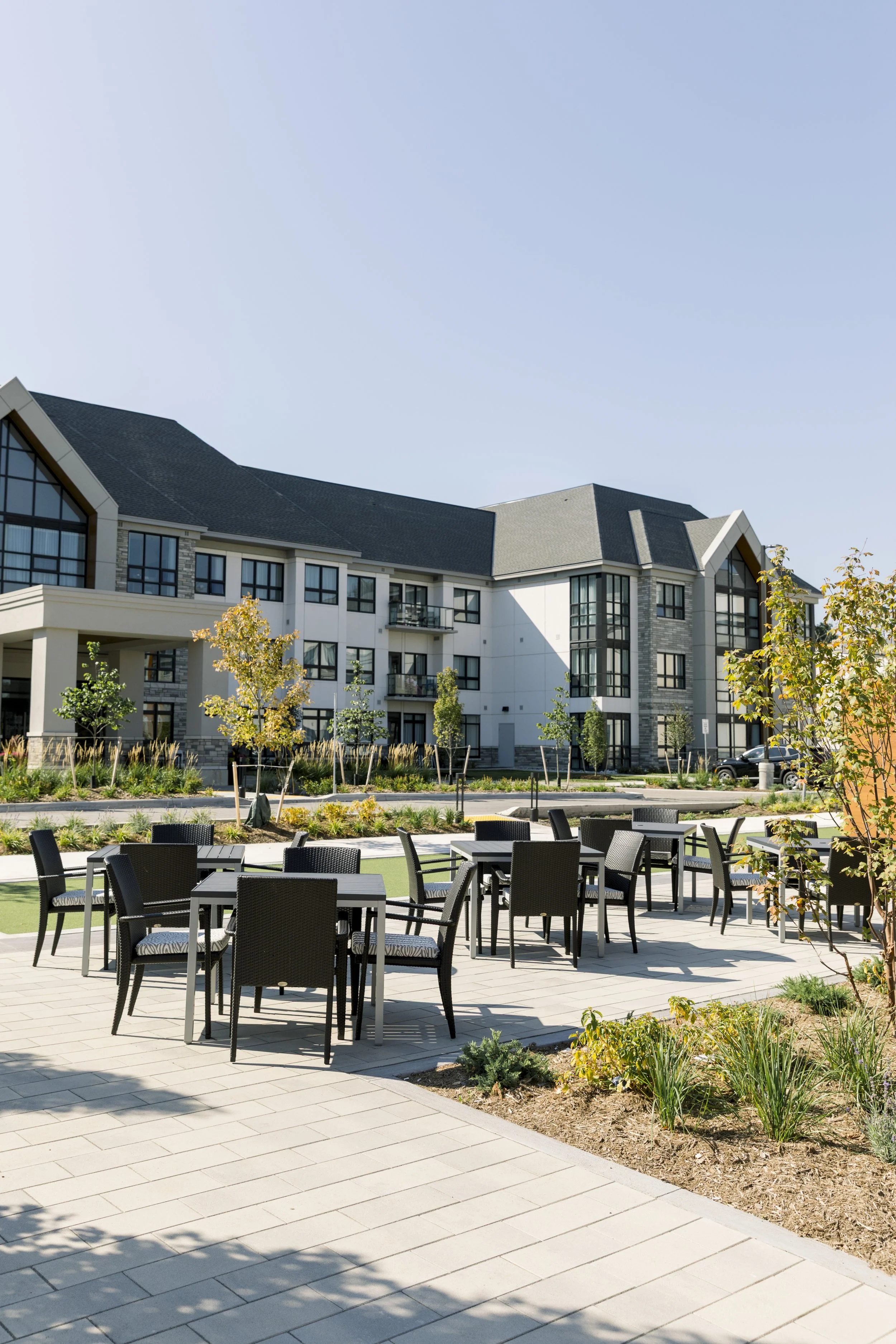 Outdoor patio with black chairs and tables, surrounded by small trees and bushes, with a modern residential building in the background under a clear blue sky.