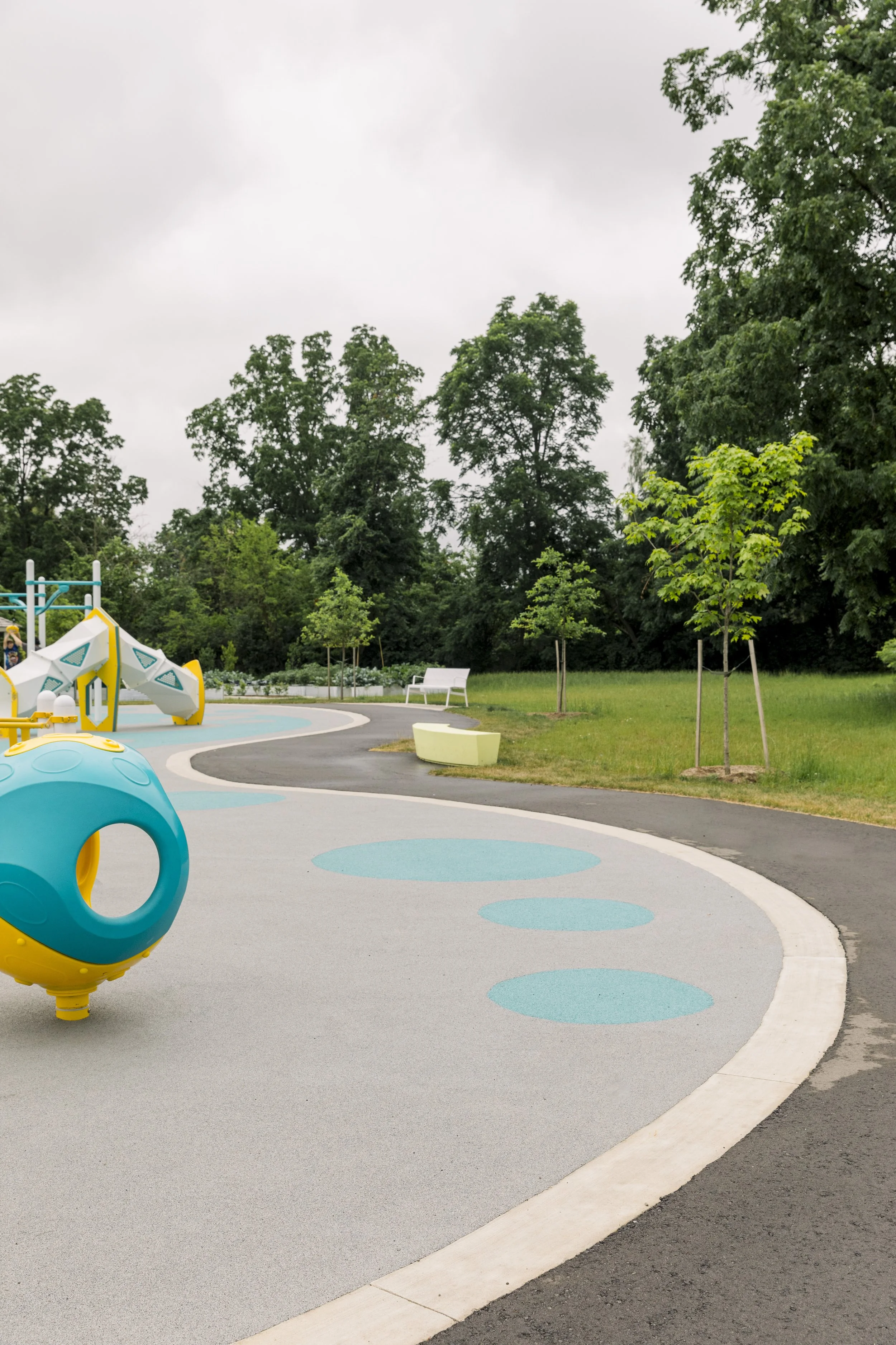 A playground with a blue and yellow climbing structure, a winding pathway, trees, grass, benches, and a cloudy sky.