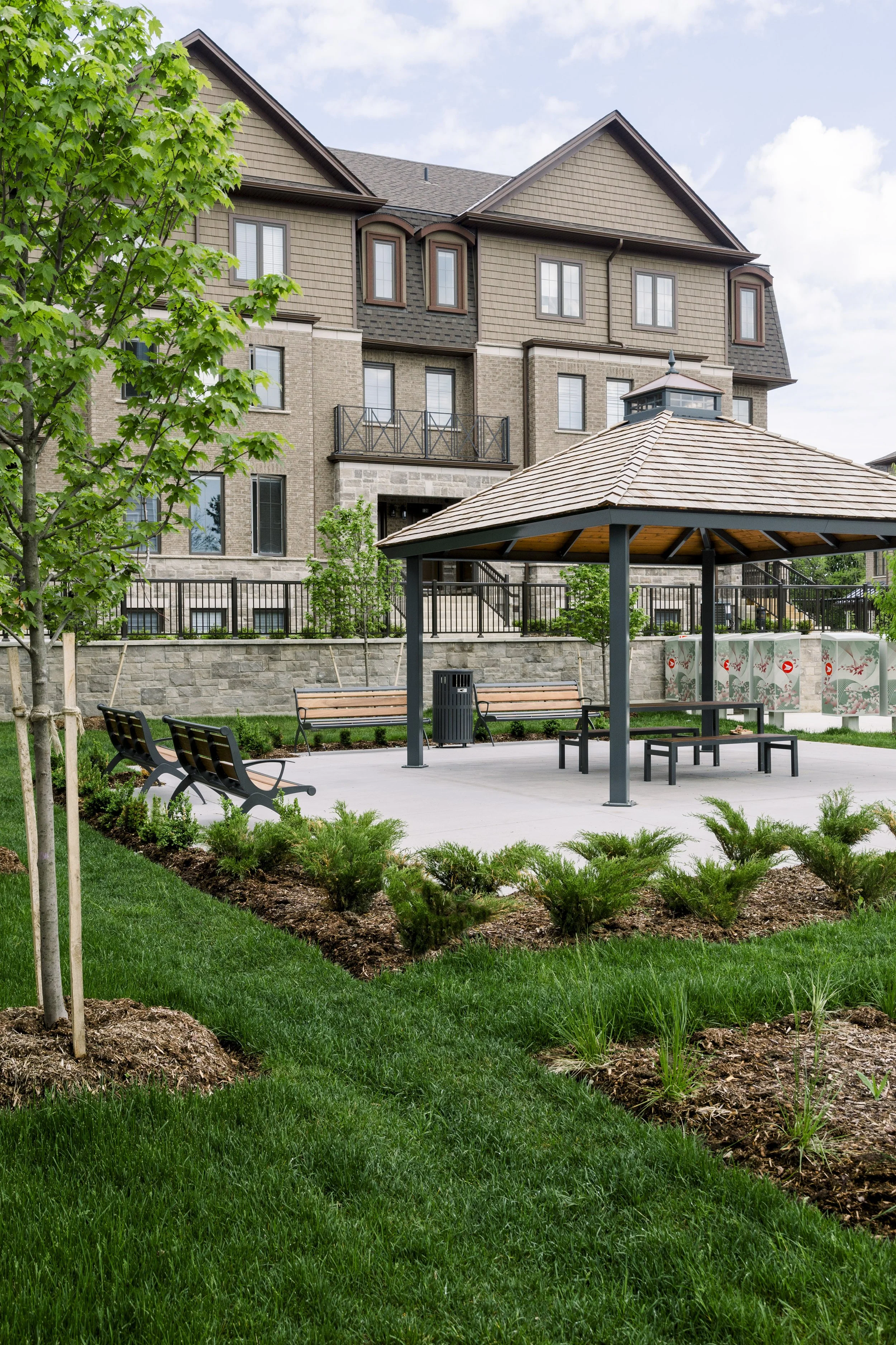 A landscaped courtyard with green grass, small plants, and young trees in front of a multi-story residential building. The courtyard features a concrete patio with a wooden picnic table under a gazebo, several benches, and a trash bin. The building h