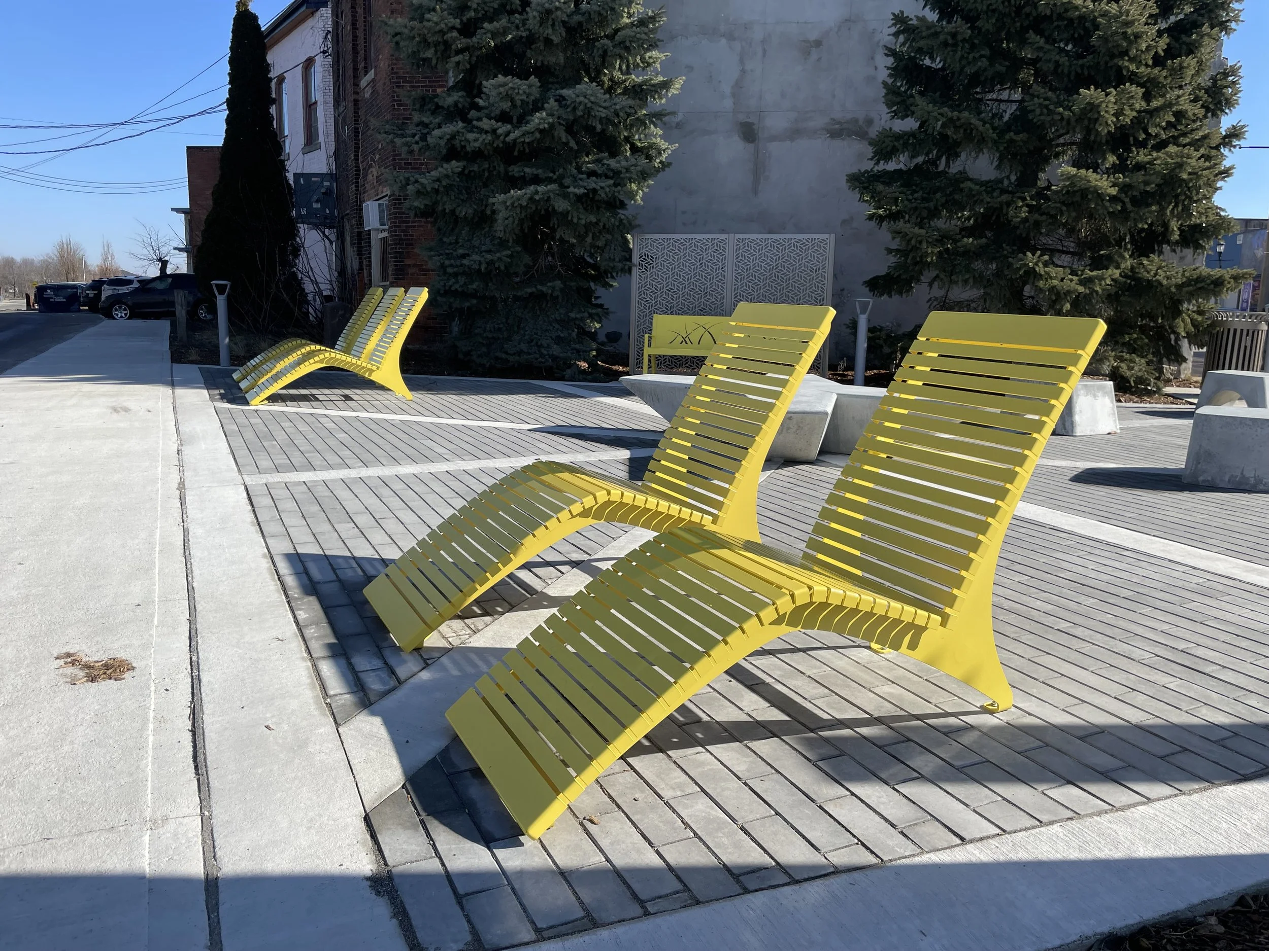 Three yellow outdoor lounge chairs are on a deserted patio, with tall trees and a building in the background.