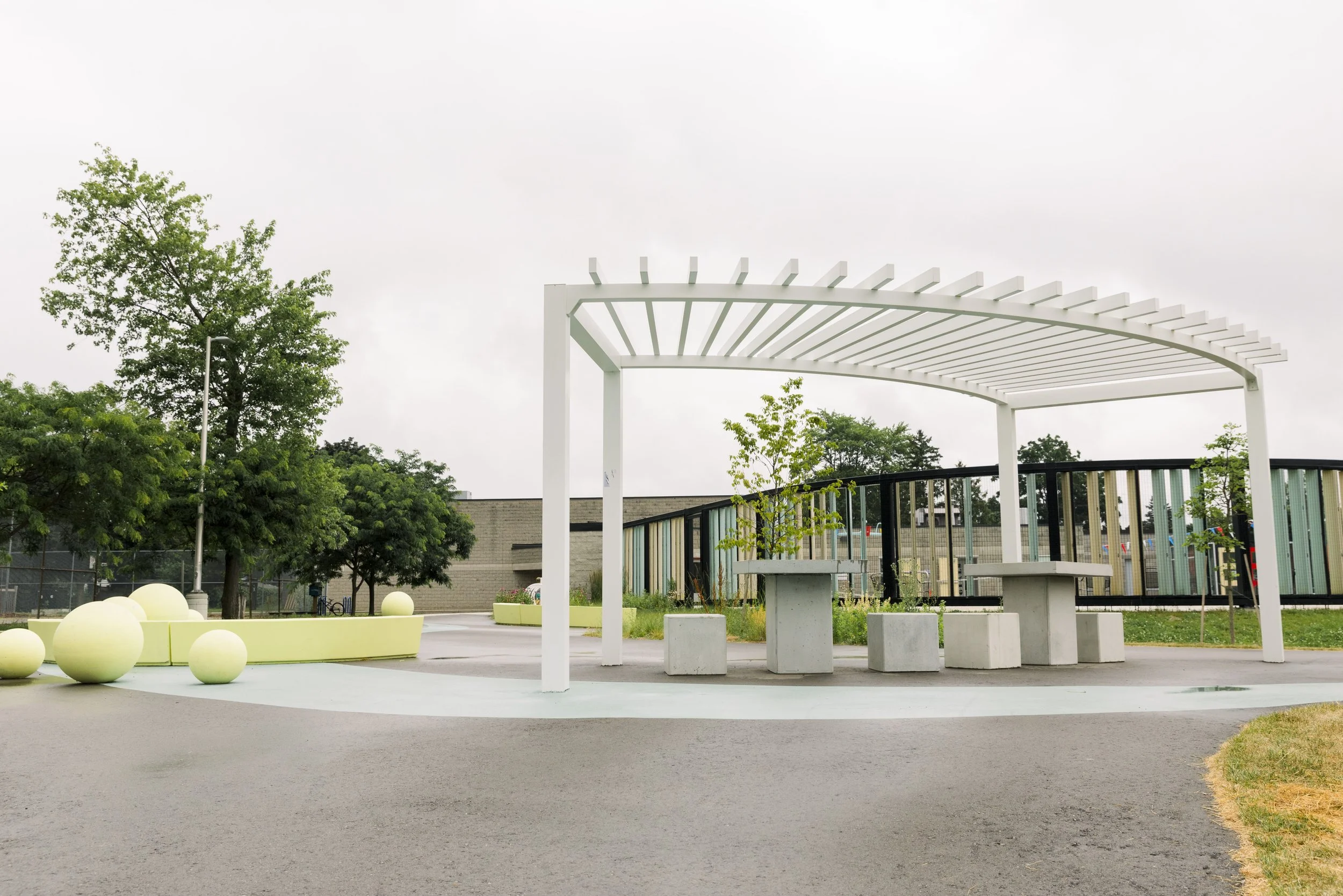 Empty outdoor park area with a white modern pergola structure, concrete tables and stools, and green trees in the background.