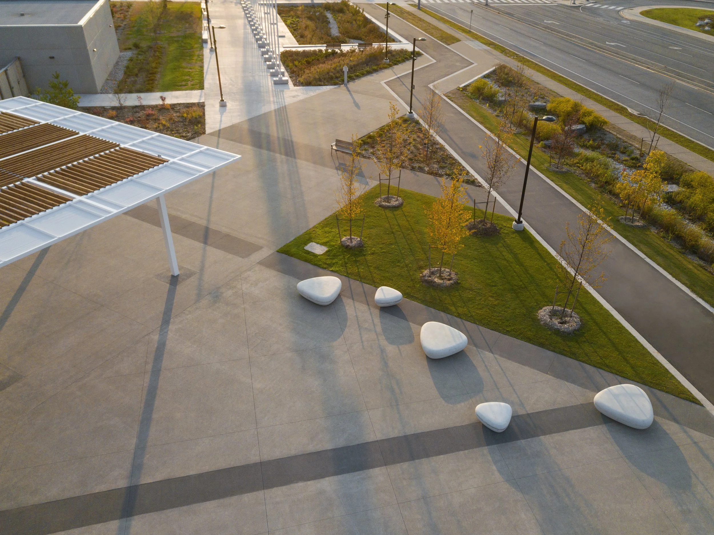 An aerial view of a landscaped outdoor area with young trees, a grassy section with stone benches, and a large concrete patio. There are lampposts along the pathways leading to a nearby road.