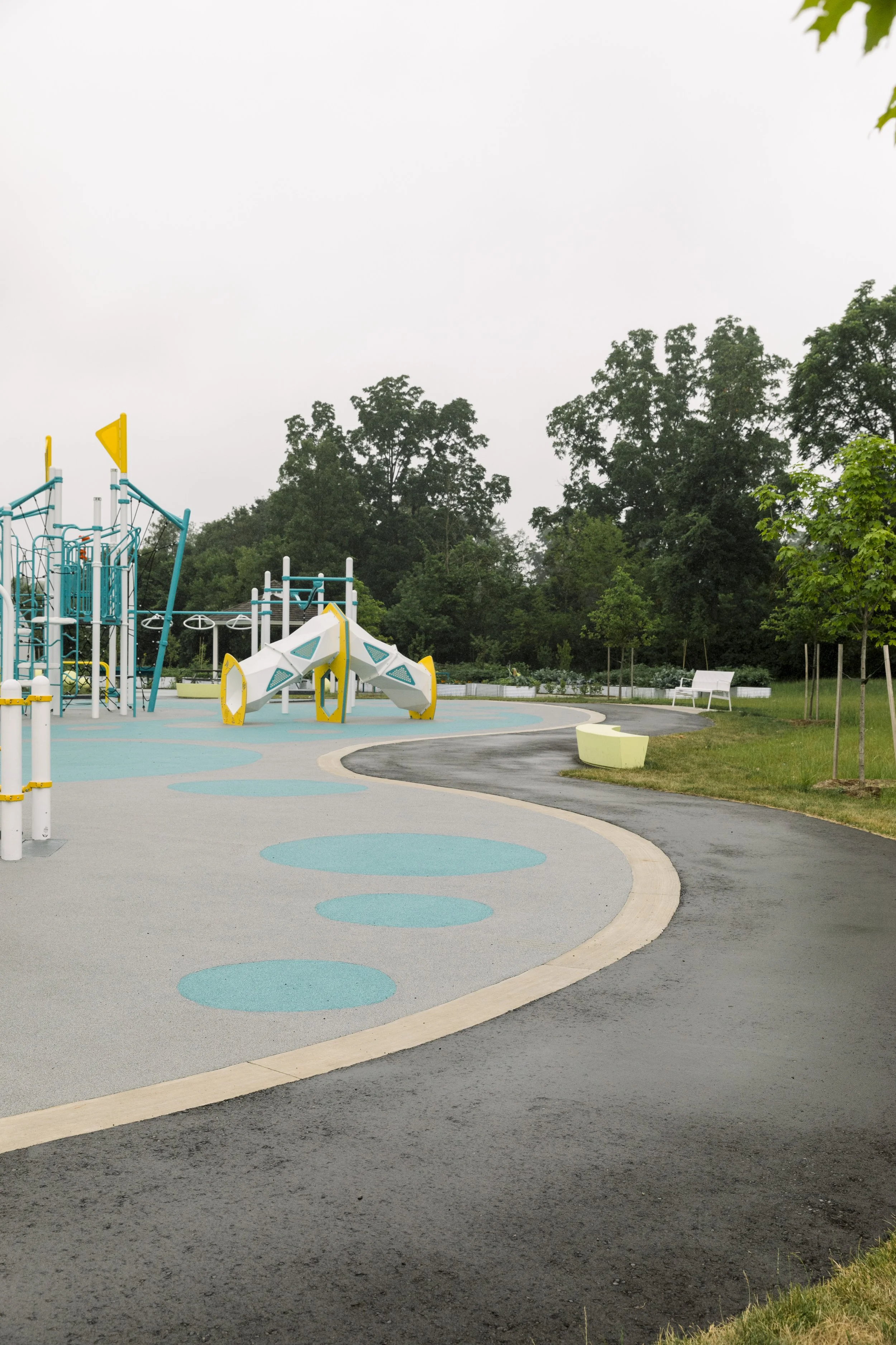 An empty playground with colorful equipment and a winding pathway surrounded by green trees and grass.