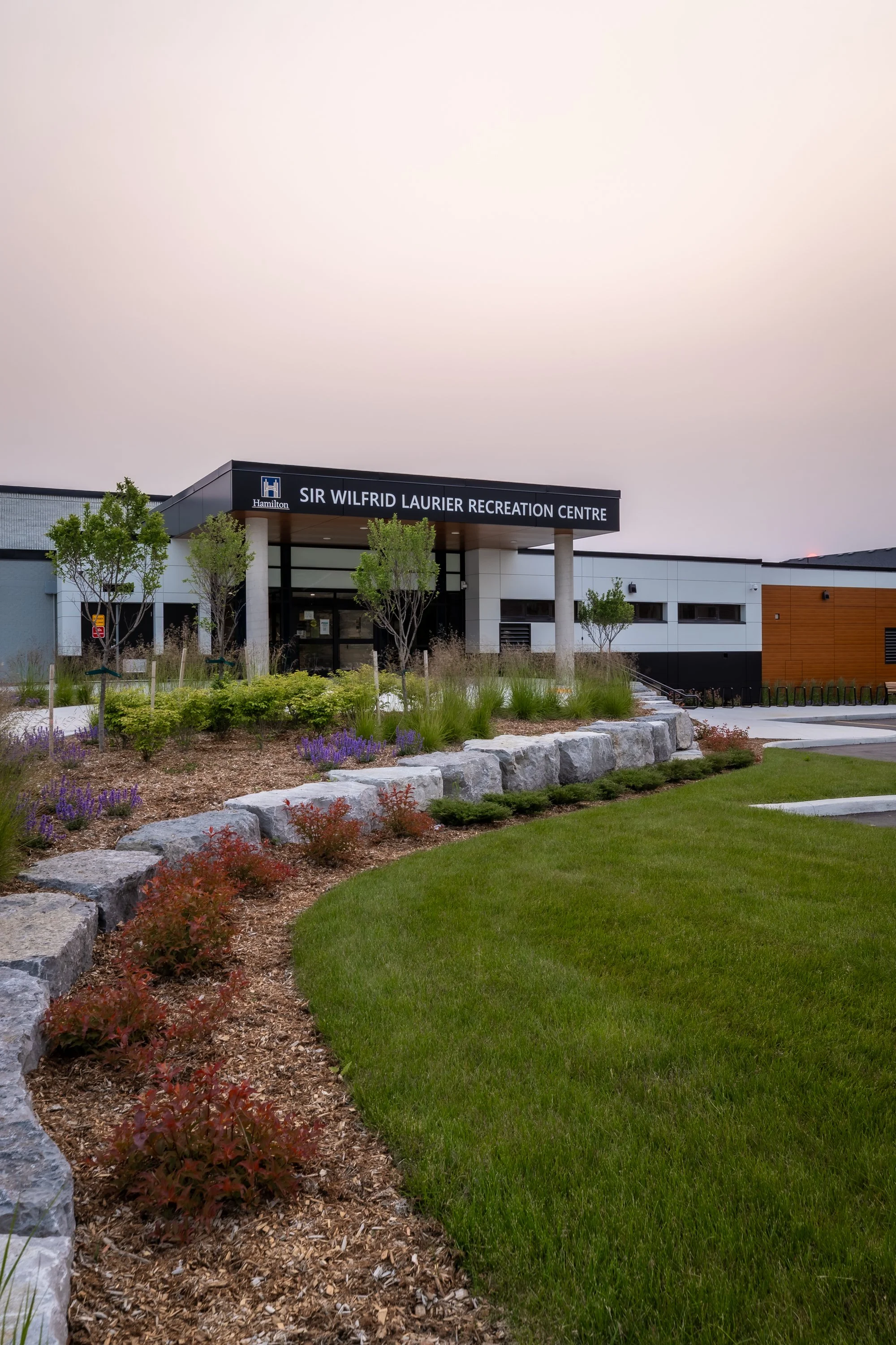 Exterior view of Sir Wilfrid Laurier Recreation Centre with landscaped garden in the foreground, trees, and a modern building facade.