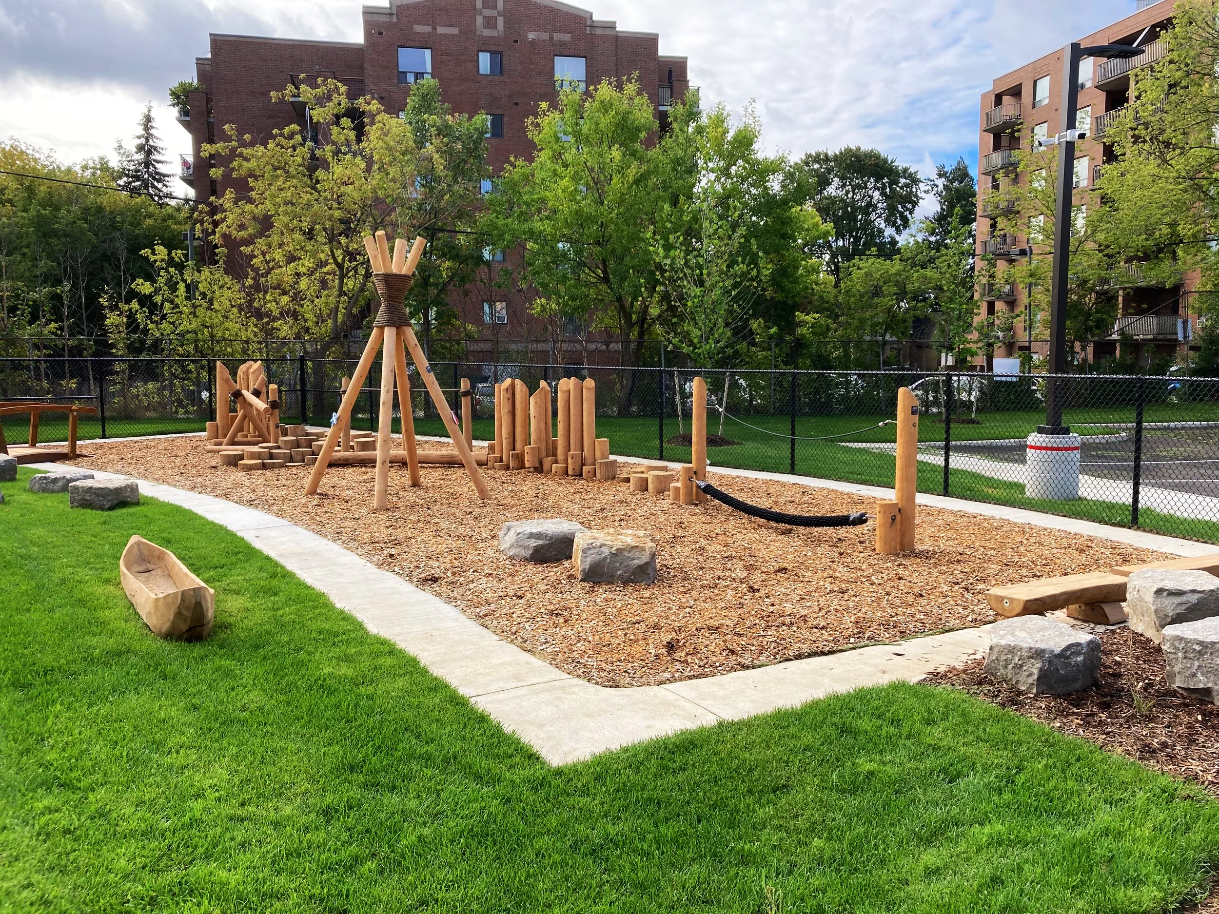 A park with a wood chip playground area enclosed by a black fence. The playground includes wooden climbing structures, a climbing log, a balance beam, and a wooden sandbox. The playground is surrounded by green grass, trees, and residential buildings in the background. The sky is partly cloudy.