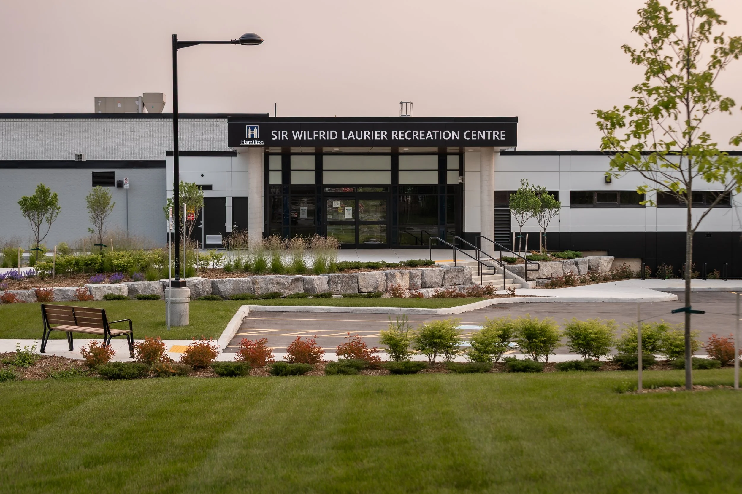 Exterior view of Sir Wilfrid Laurier Recreation Centre with landscaping, pathway, bench, and parking lot in front.