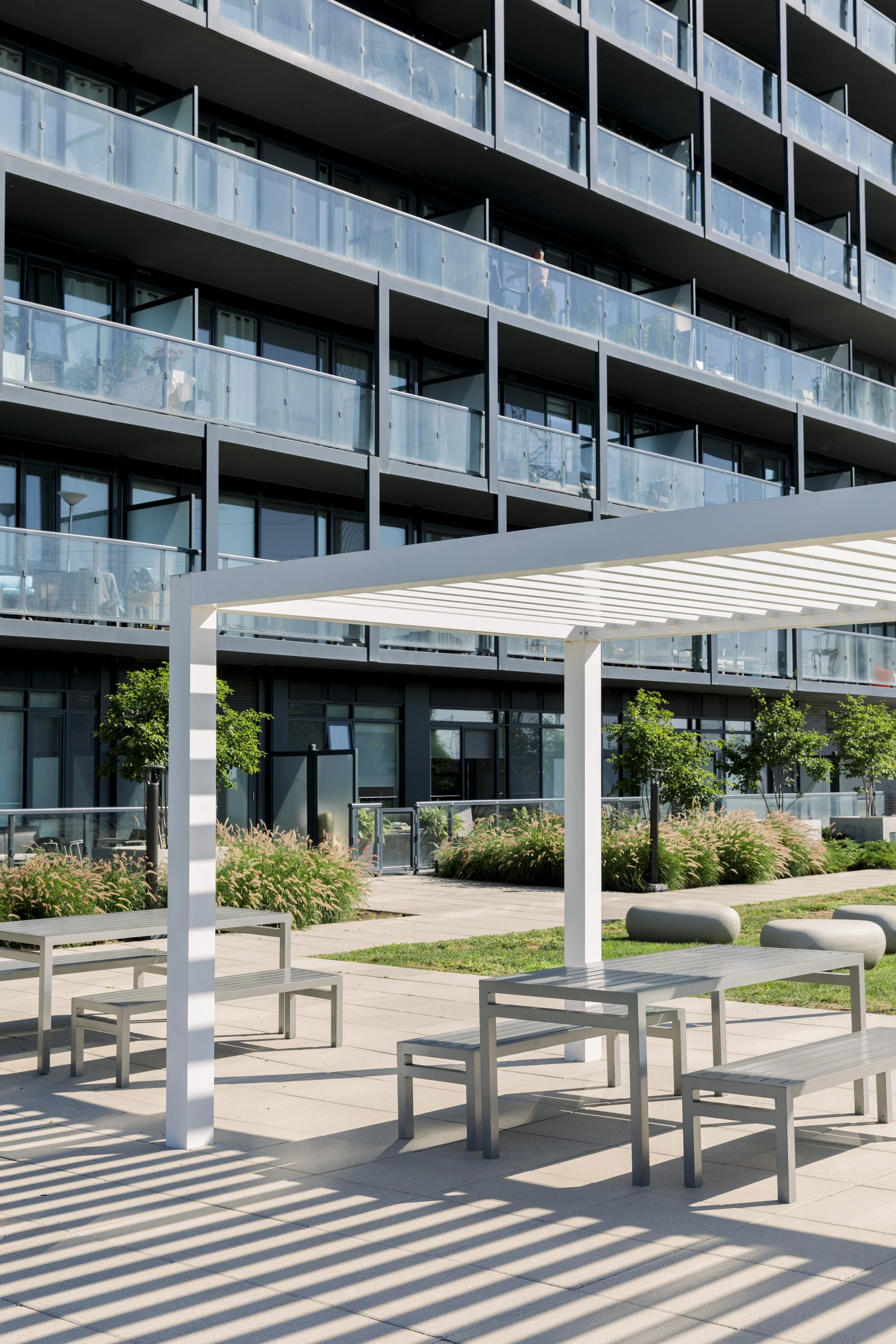 Modern apartment building with balconies, outdoor seating area with benches and tables, shaded by a white pergola, surrounded by landscaped greenery.