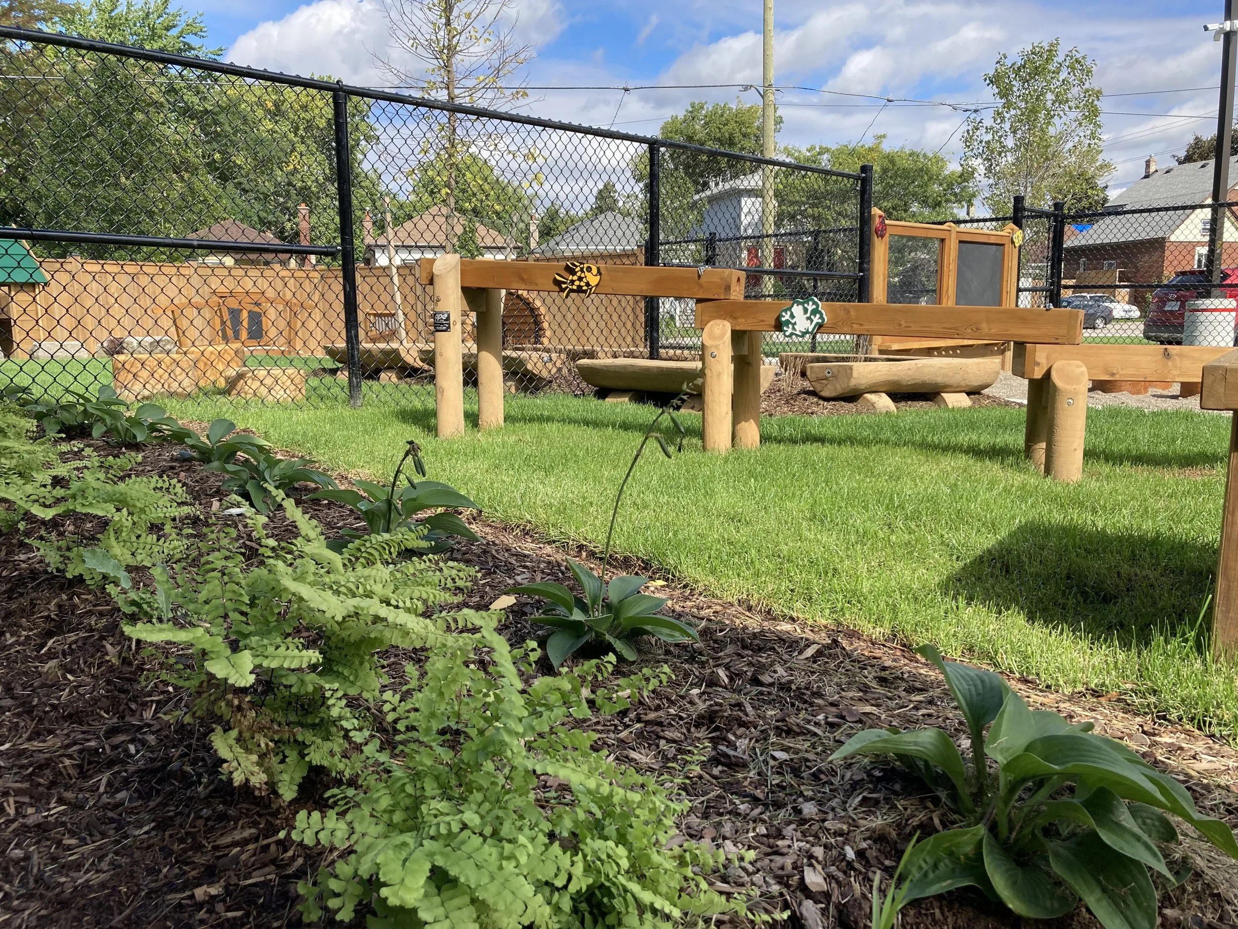 A fenced outdoor area with a grassy yard, wooden pipe structures, and a small garden bed in the foreground. Neighborhood houses are visible in the background under a partly cloudy sky.