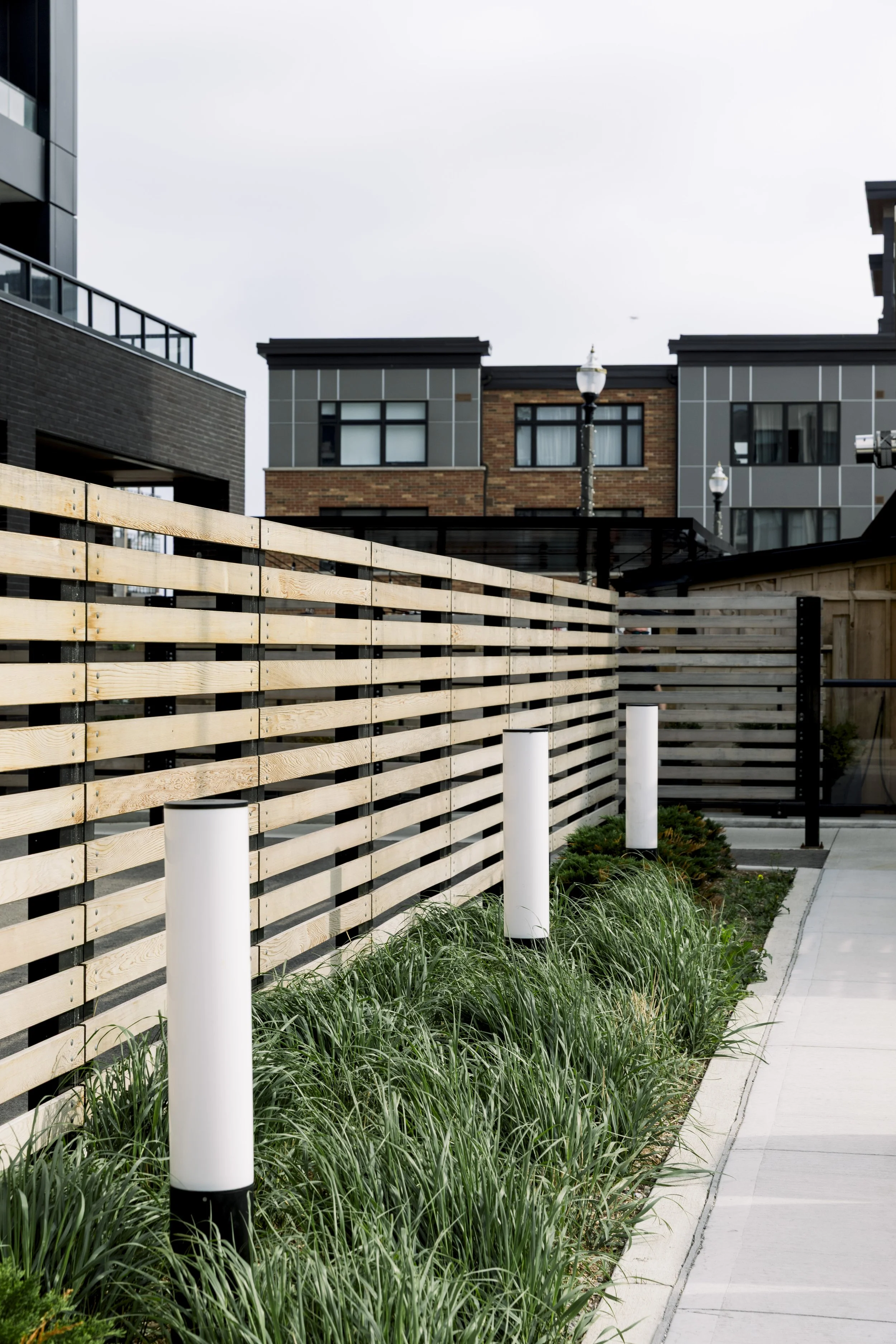 Modern outdoor scene with a wooden slat fence, landscaped greenery, and a sidewalk in an urban area with apartment buildings in the background.