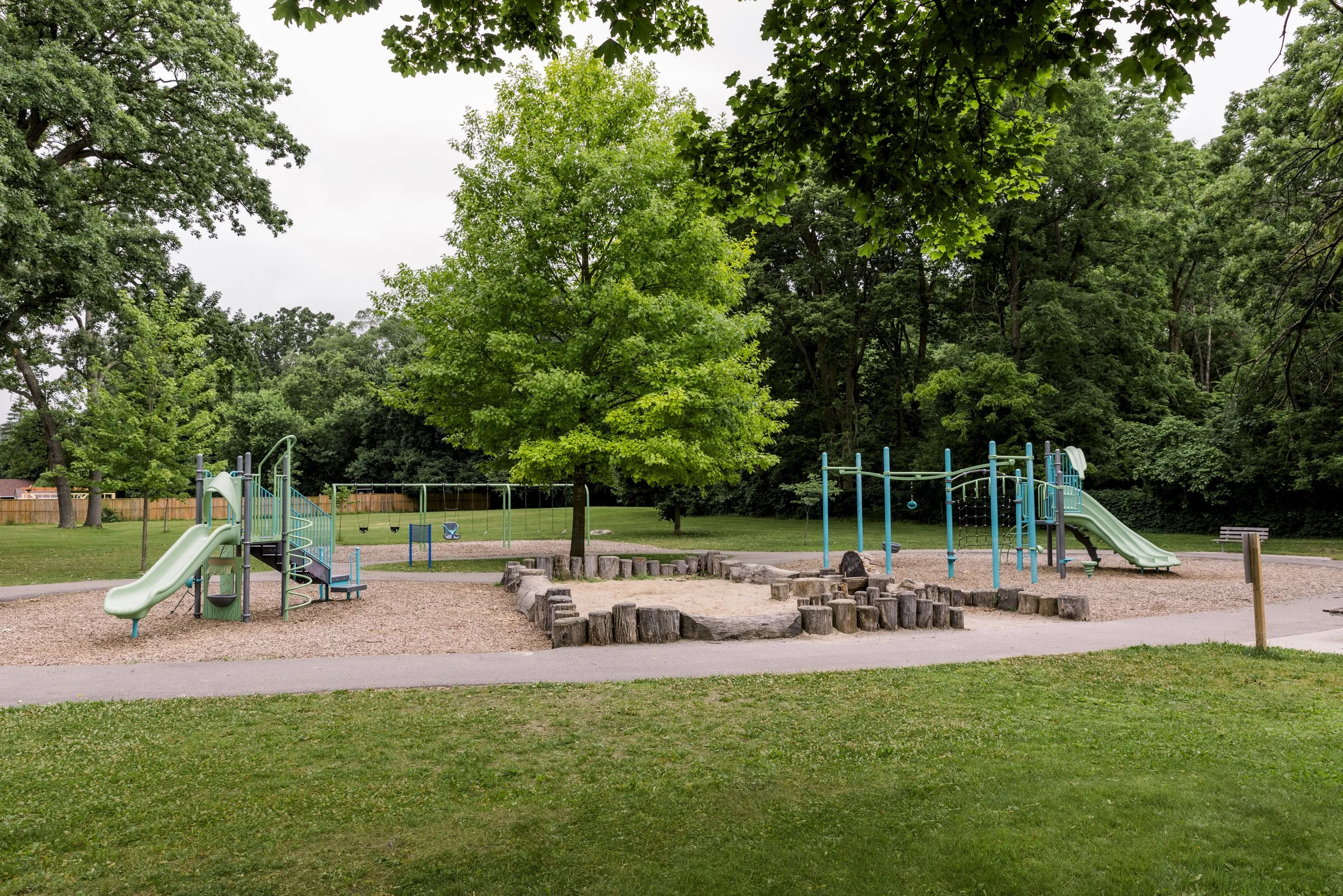 Empty playground with slides, swings, climbing bars, sand area, surrounded by trees and grass.