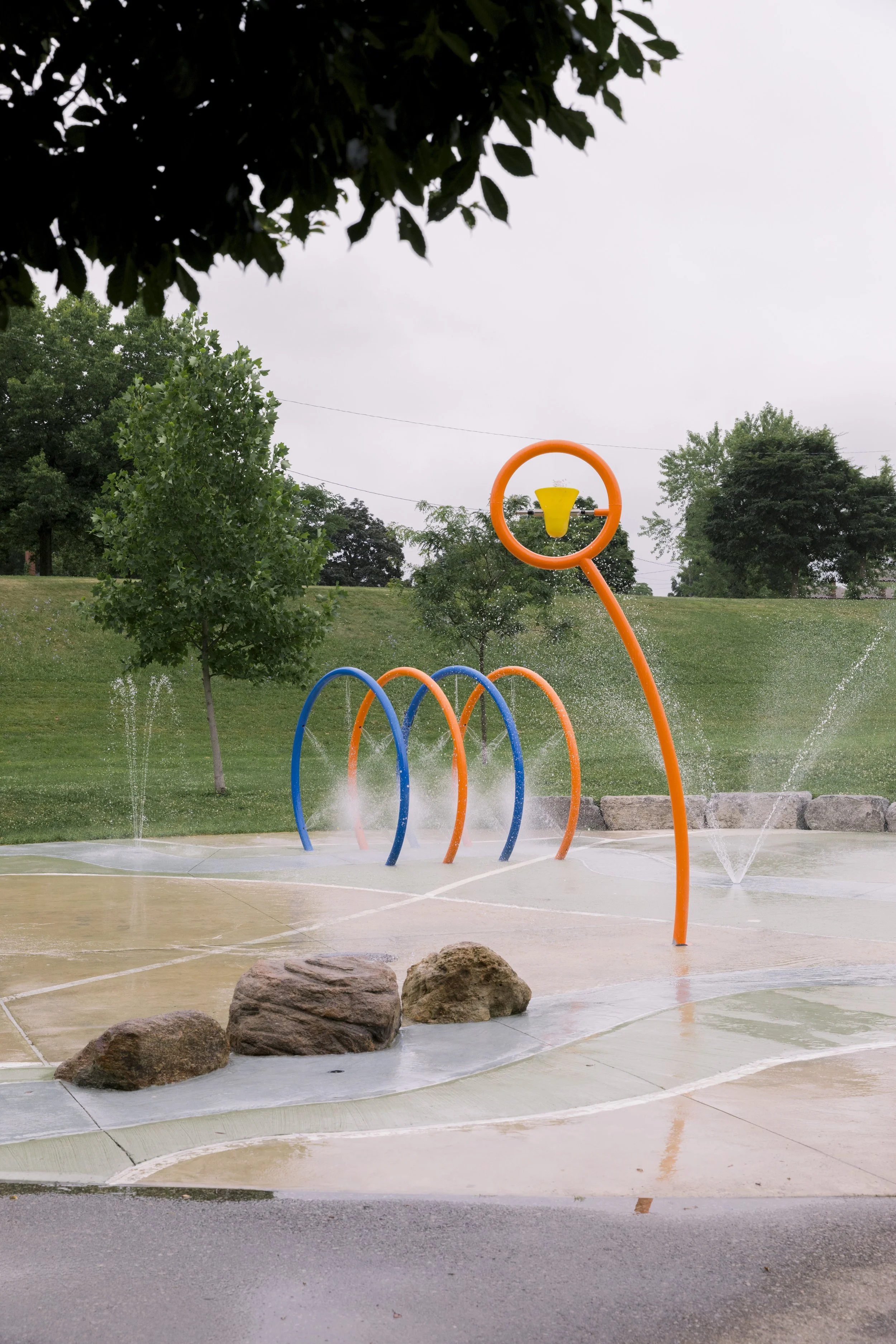 Colorful outdoor splash pad with rings and water sprays, surrounded by rocks and green trees on overcast day.