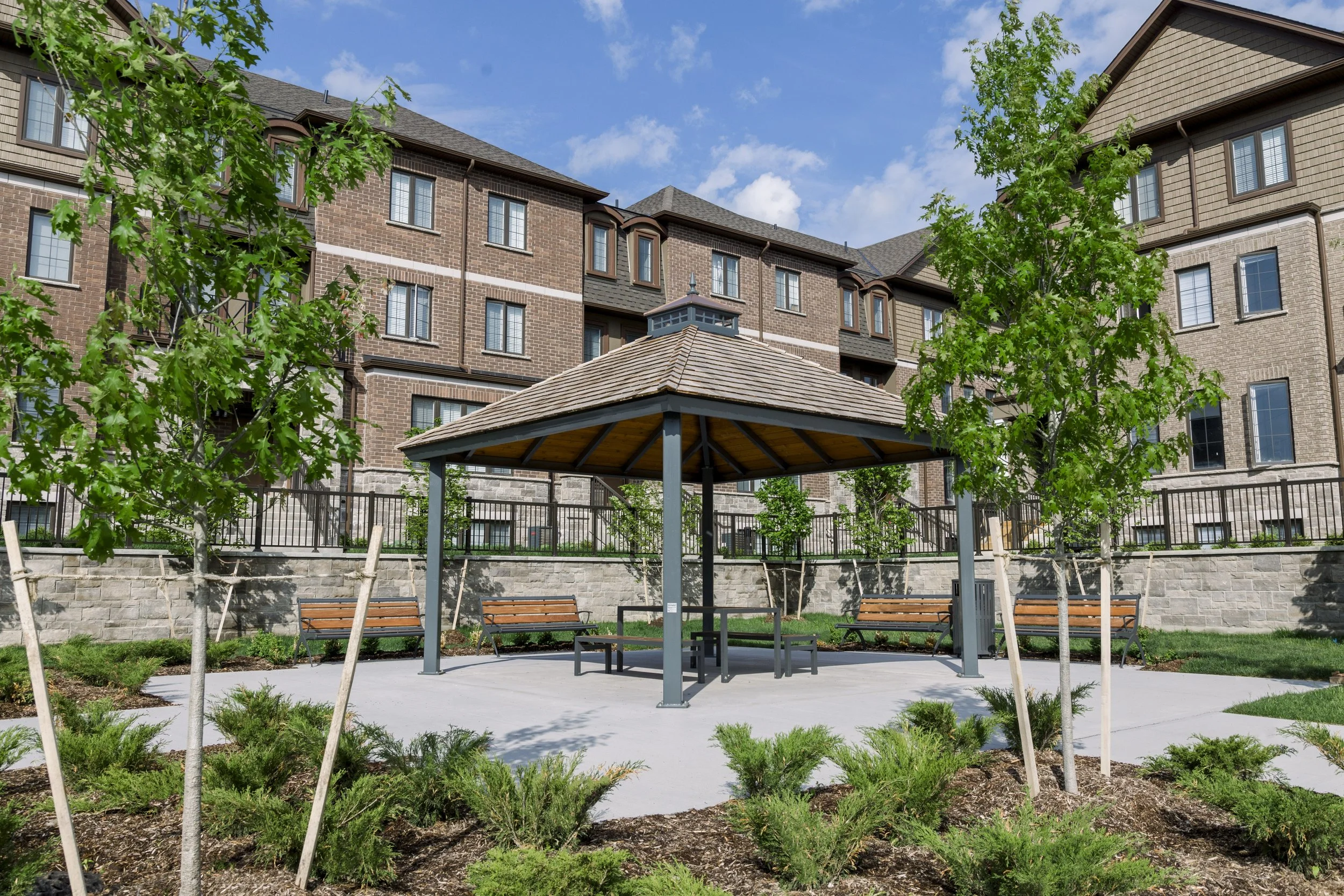 A small park with a gazebo, surrounding benches, trees, and shrubbery in front of multi-story apartment buildings under a blue sky with clouds.