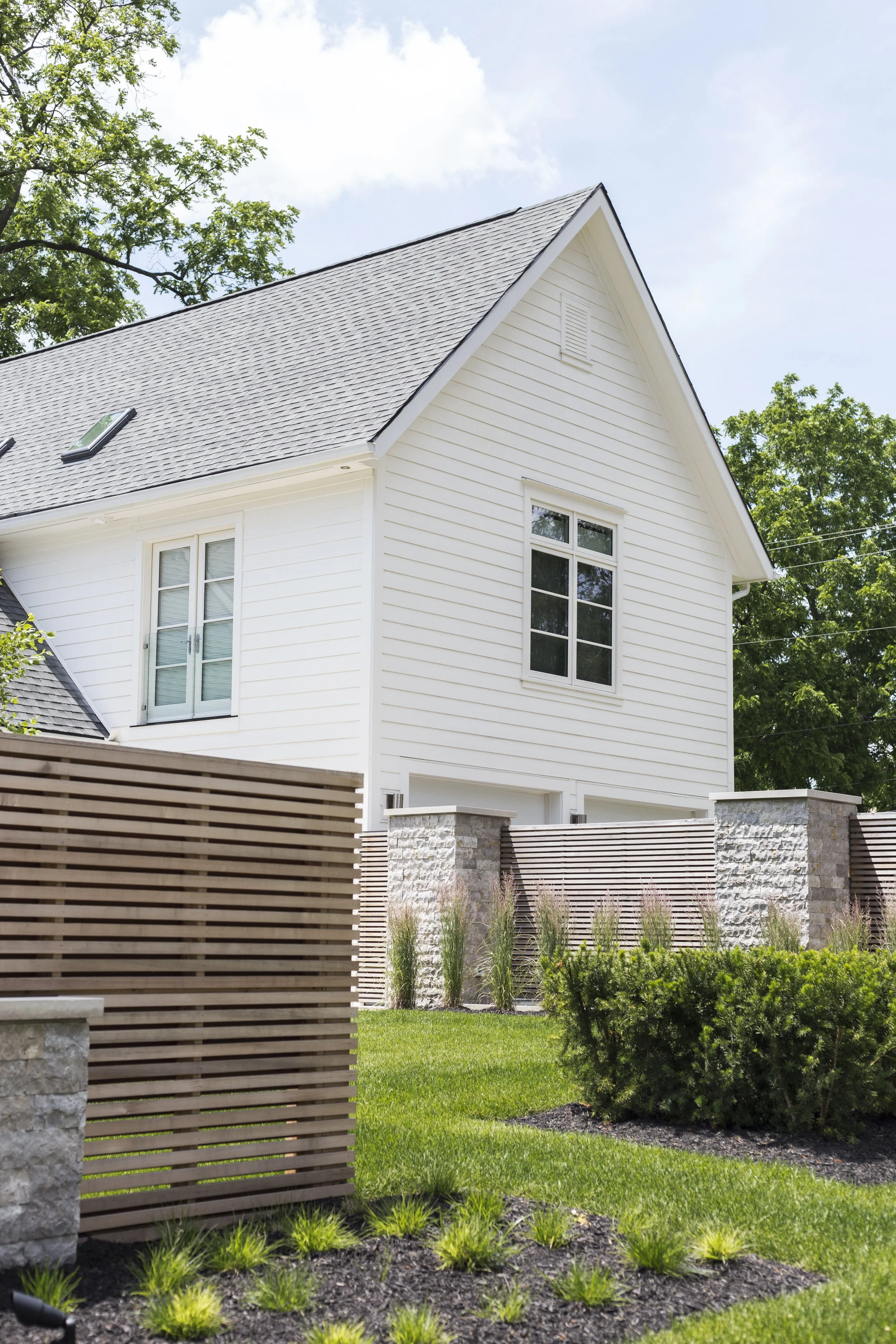 A white two-story house with a steep gray roof, large windows, and a fenced yard with green grass and shrubs.