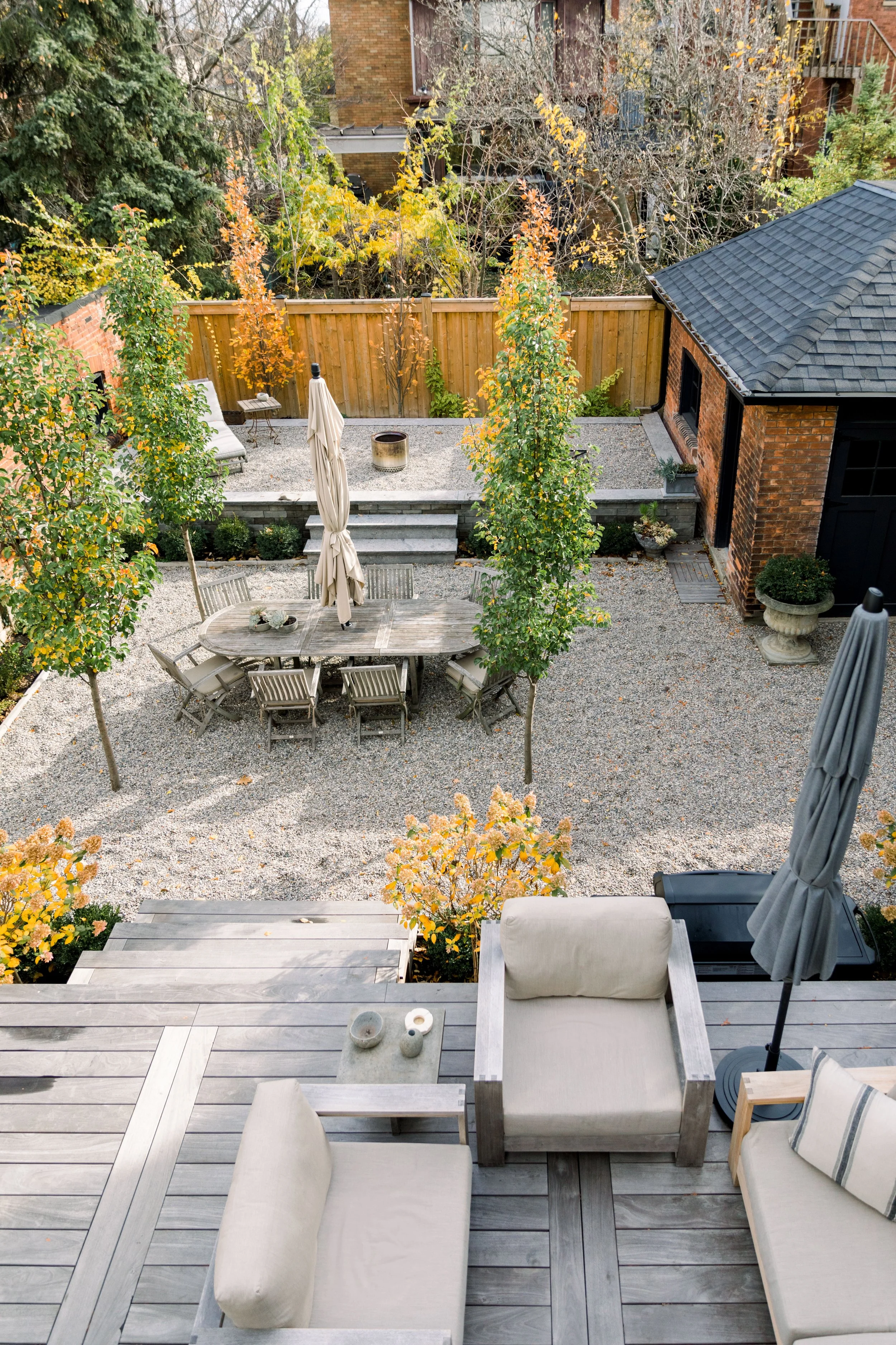 A backyard patio with outdoor furniture, trees with fall-colored leaves, a gravel area, and a wooden fence.