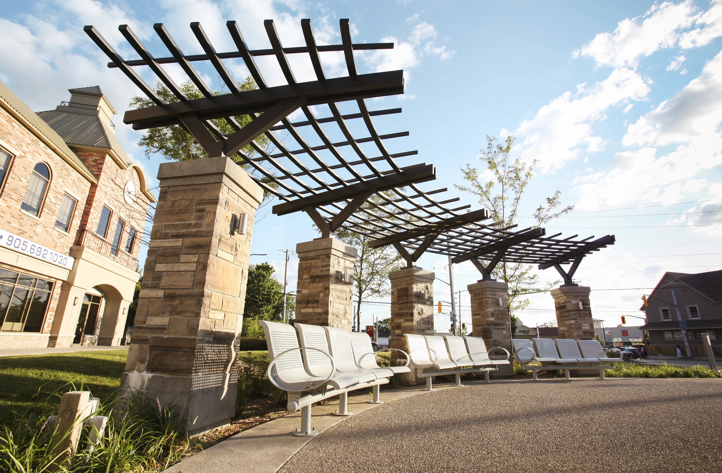 Outdoor seating area with multiple white benches under a wooden pergola supported by stone columns, with buildings and a busy street in the background, and clear sky with scattered clouds.