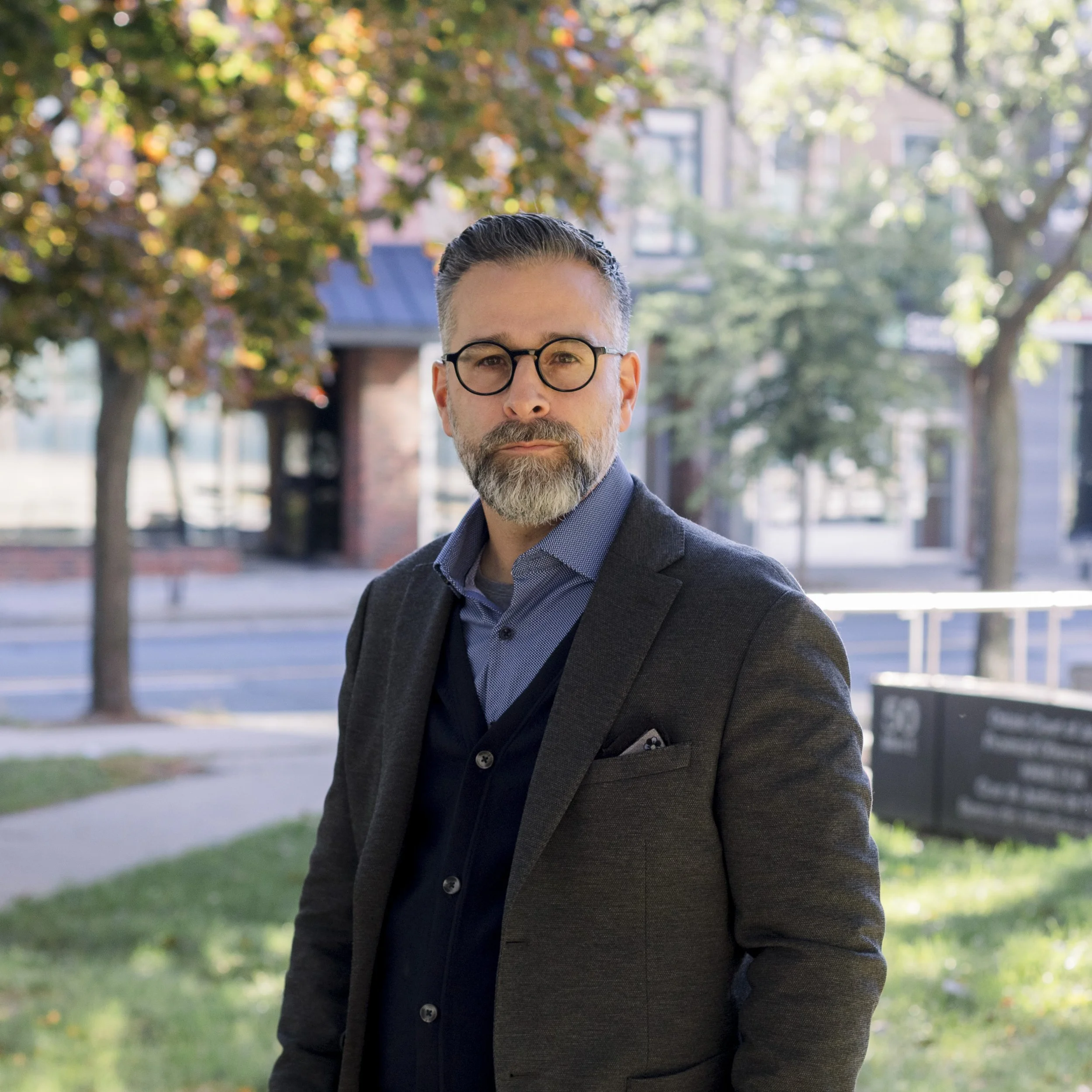A middle-aged man with glasses, grey hair, and a beard stands outdoors on a sunny day, wearing a dark blazer over a blue shirt with trees and buildings in the background.