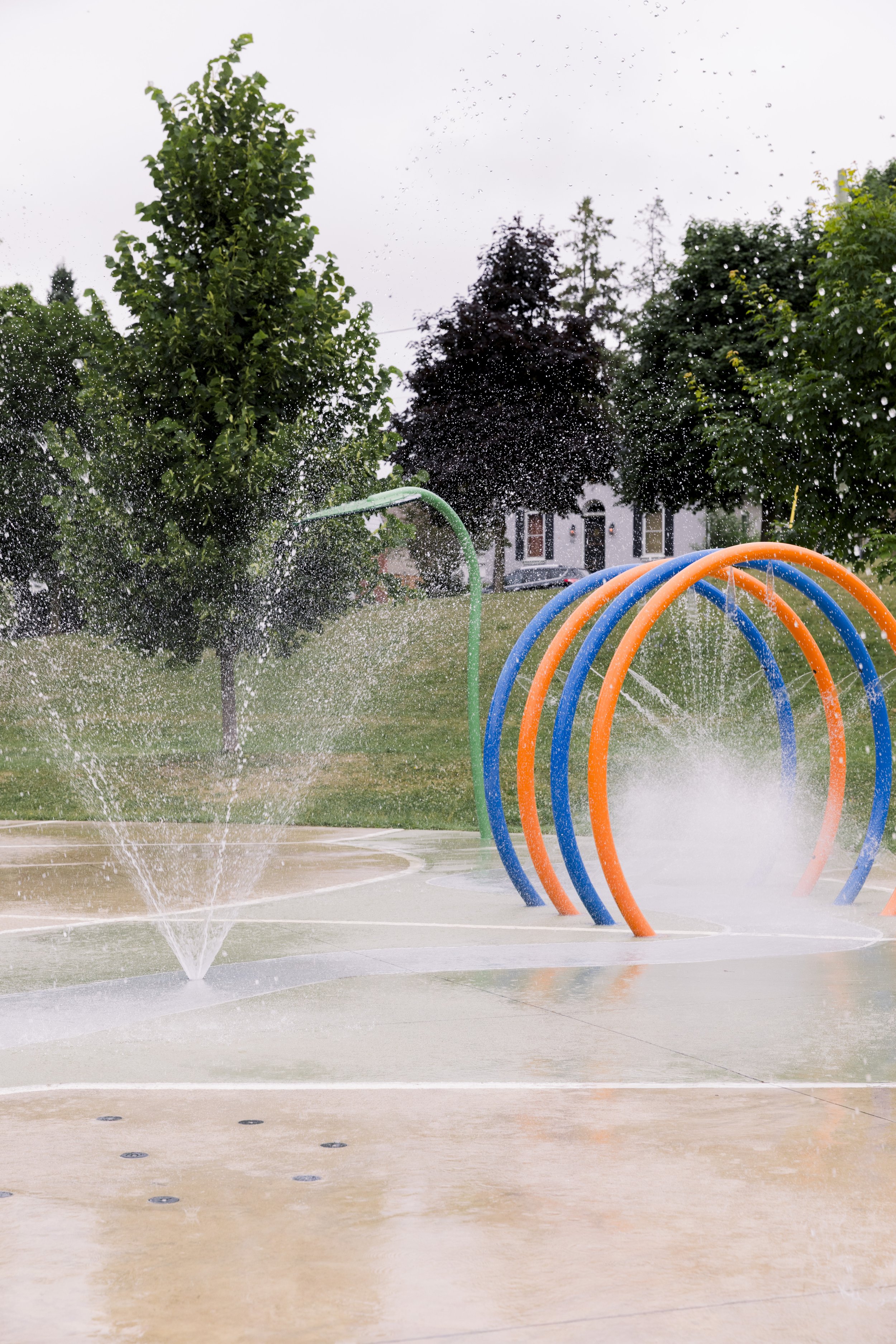 Colorful playground water fountain with spray nozzles and arches, surrounded by green trees and grass, with a house in the background.