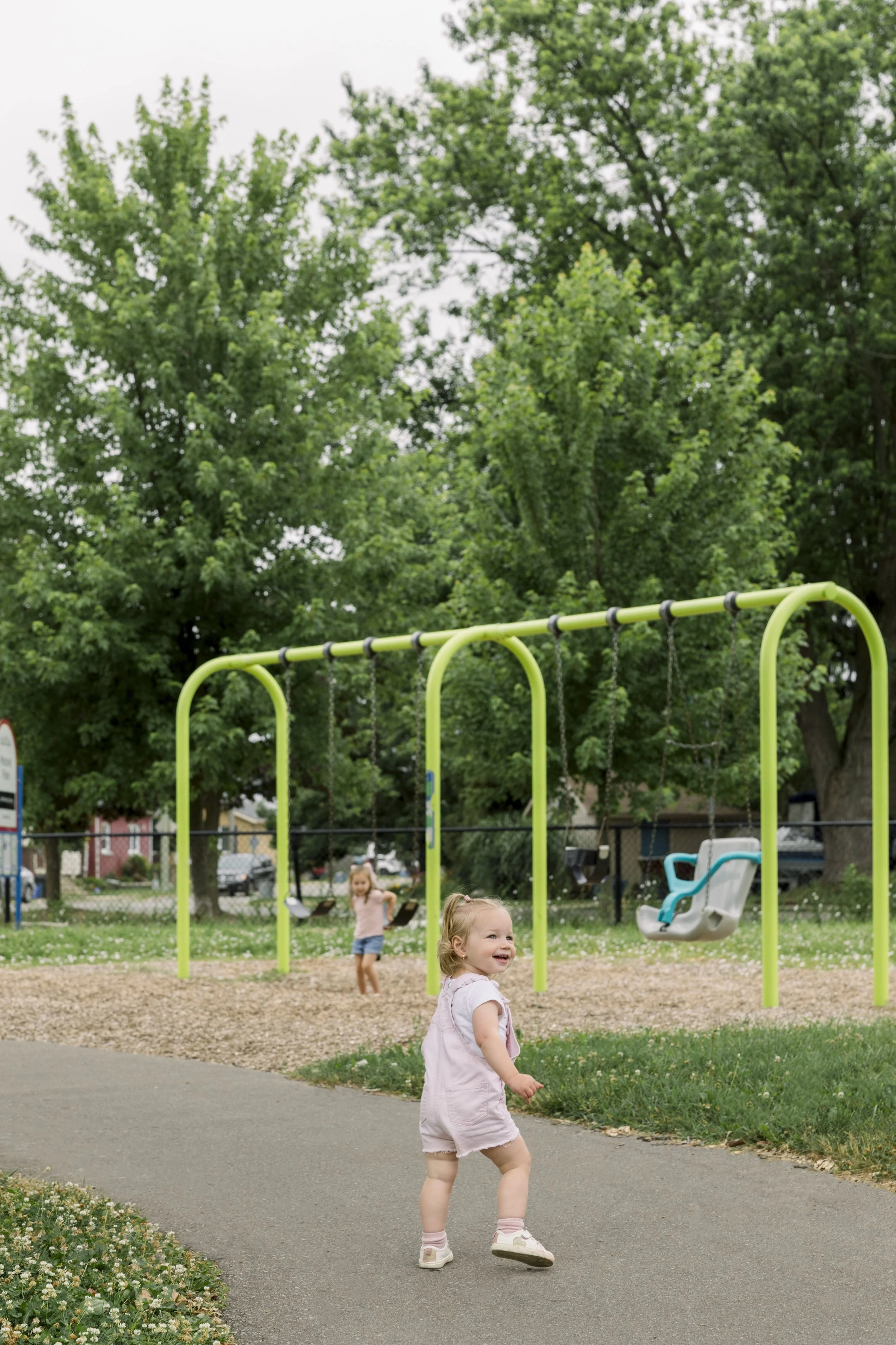Children playing on swings and walking in a park with trees and a paved path.
