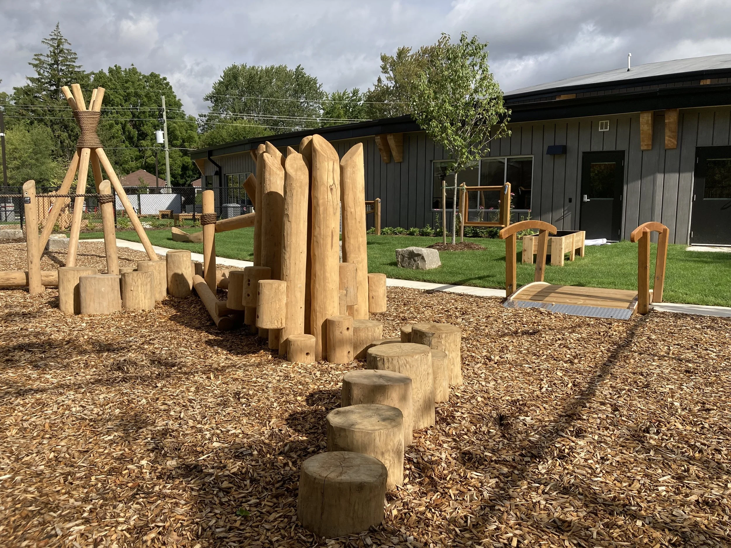 Wooden outdoor playground with logs, stepping stones, a slide, and a teepee structure. Building with dark gray exterior walls is visible in the background, surrounded by green grass, a tree, and a cloudy sky.