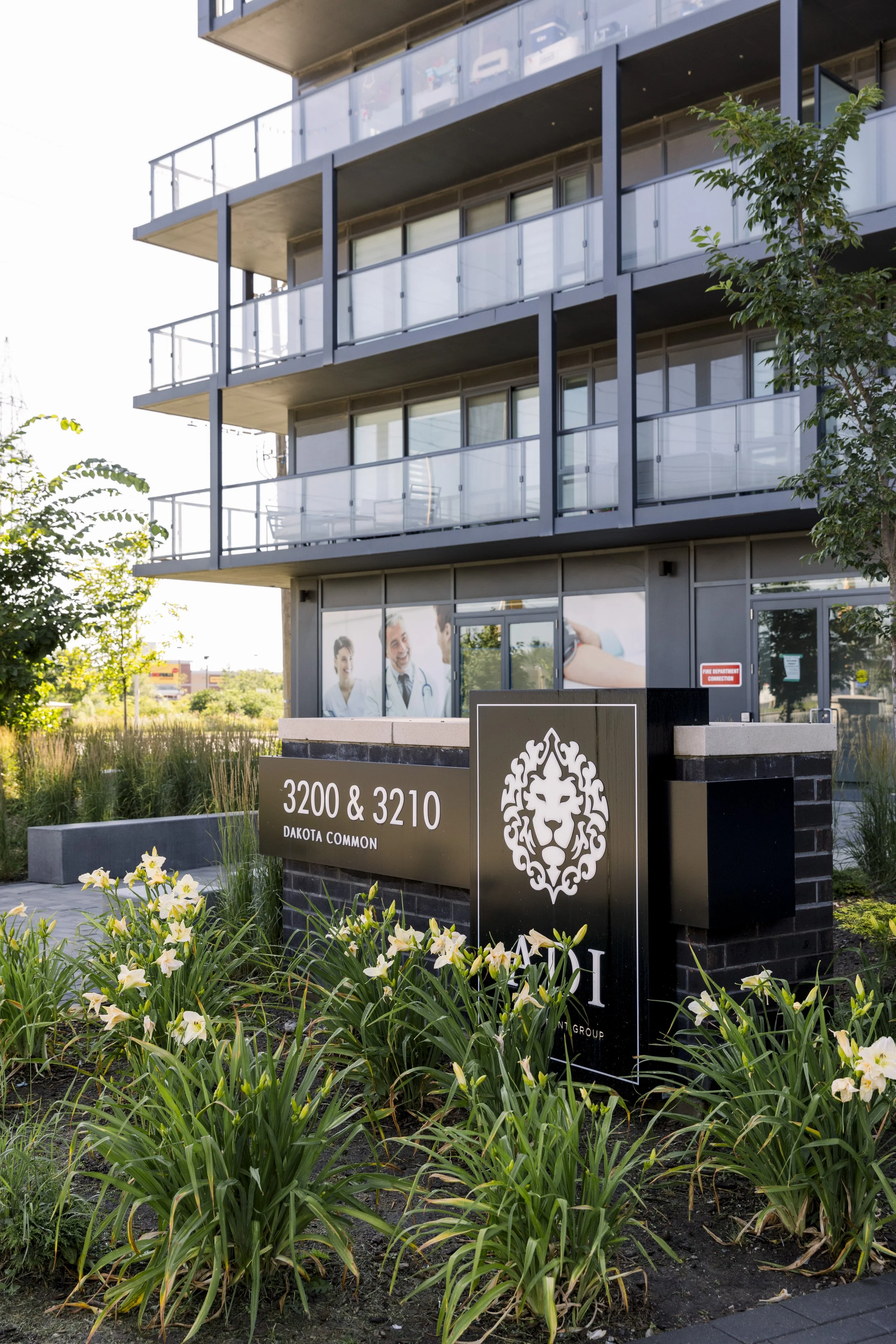 Exterior of a modern multi-story building with glass balconies, part of a medical or healthcare facility named DAKOTA COMMON, with flower beds and a sign displaying the address 3200 & 3210.